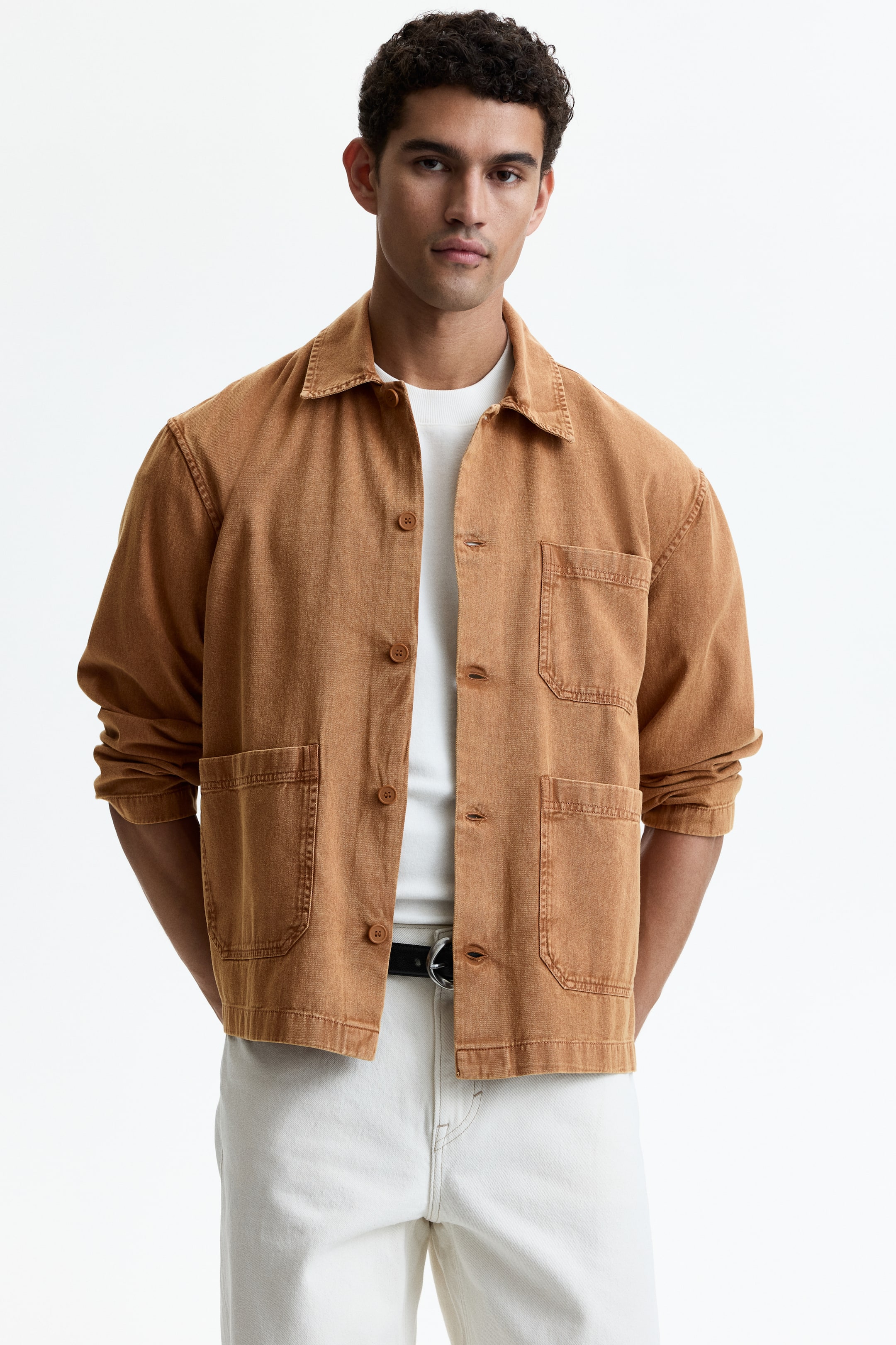 View larger image: The model is showcasing a rust-hued cotton twill overshirt, designed with a collar and button-front closure. It features two patch pockets at the front, a patch chest pocket, long sleeves, and a straight-cut hem, worn open over a white t-shirt.