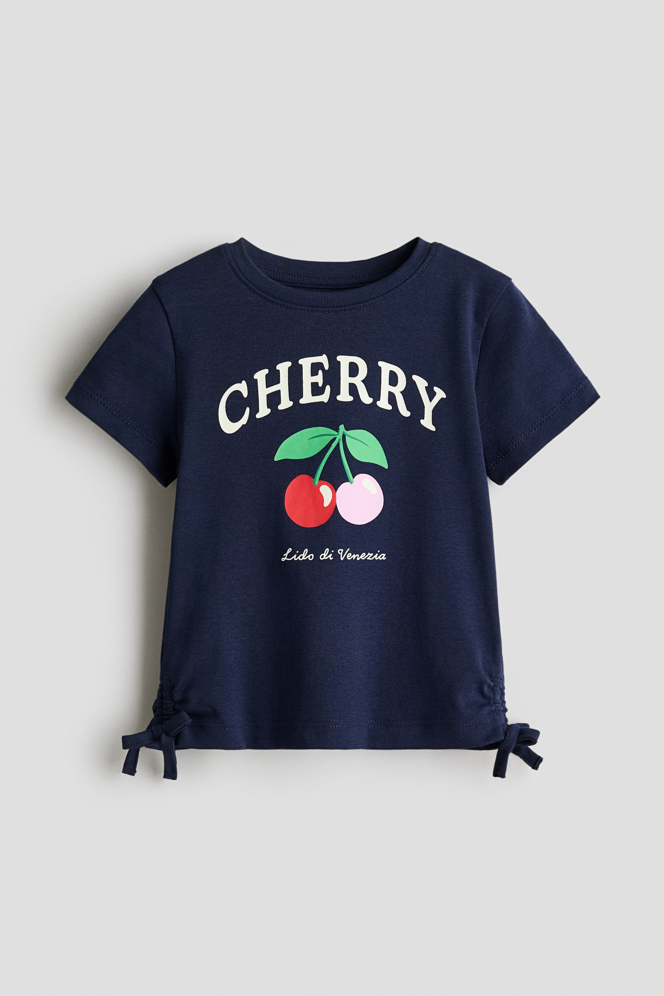 Xem ảnh lớn hơn: Một chiếc áo thun màu xanh đậm đang nằm phẳng, hướng về phía trước, với cổ tròn, tay ngắn và các đường gân bên được cột thành nơ ở gấu áo. Mặt trước hiển thị một hình in đồ họa với từ "CHERRY" bằng chữ in hoa màu trắng ngà, được đặt phía trên hình vẽ của hai trái cherry?một đỏ, một hồng nhạt, với cuống và lá màu xanh?và "Lido di Venezia" bằng chữ viết trắng bên dưới.