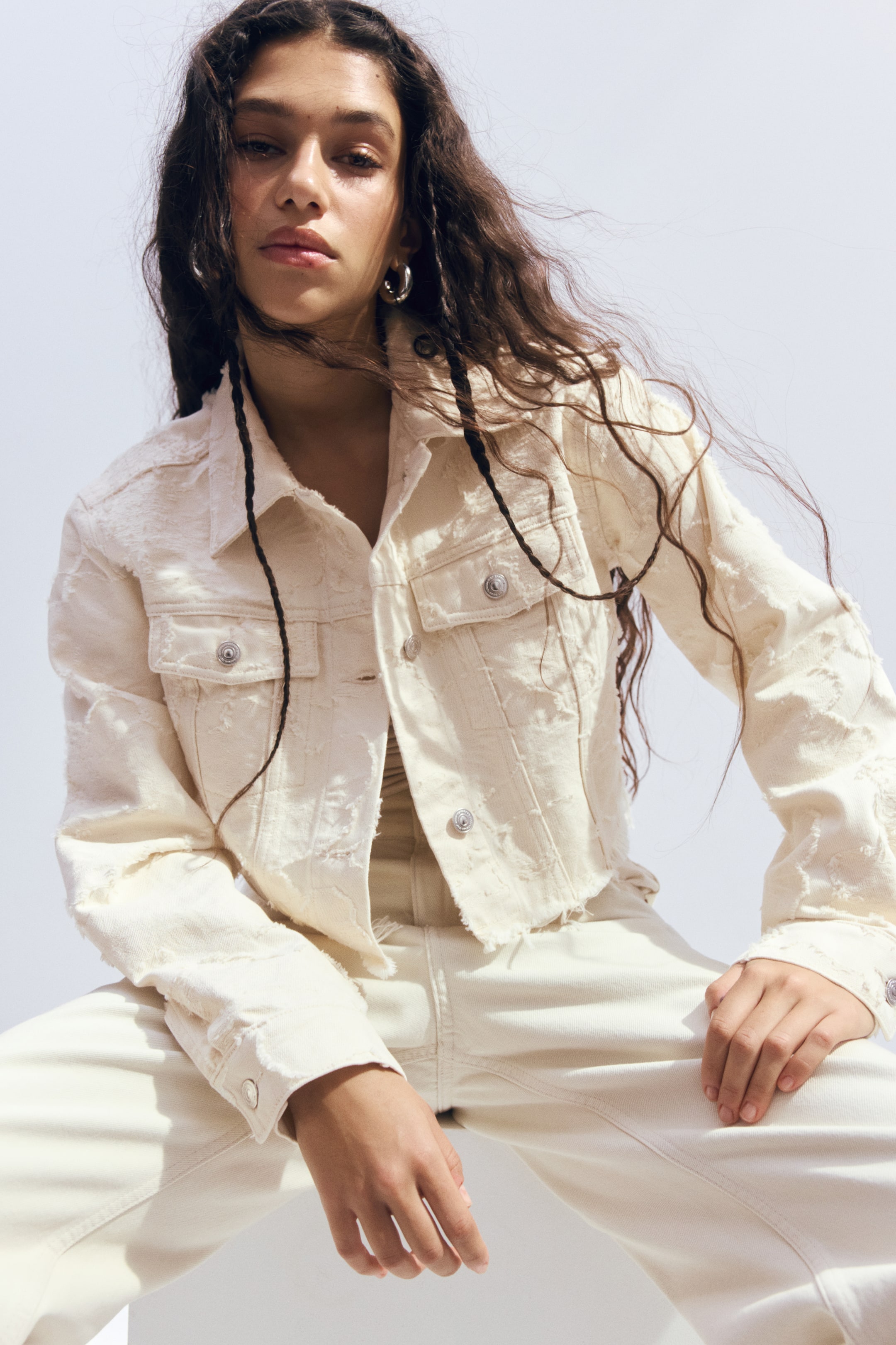 Agrandir l'image: Une veste en jean raccourcie de couleur crème est portée, dotée d'un col pointu et d'une fermeture boutonnée à l'avant avec des boutons argentés. Elle présente deux poches poitrine à rabat avec fermetures à boutons, des manches longues avec poignets boutonnés, et une apparence usée avec des détails effilochés et des patchs texturés sur l'ensemble.