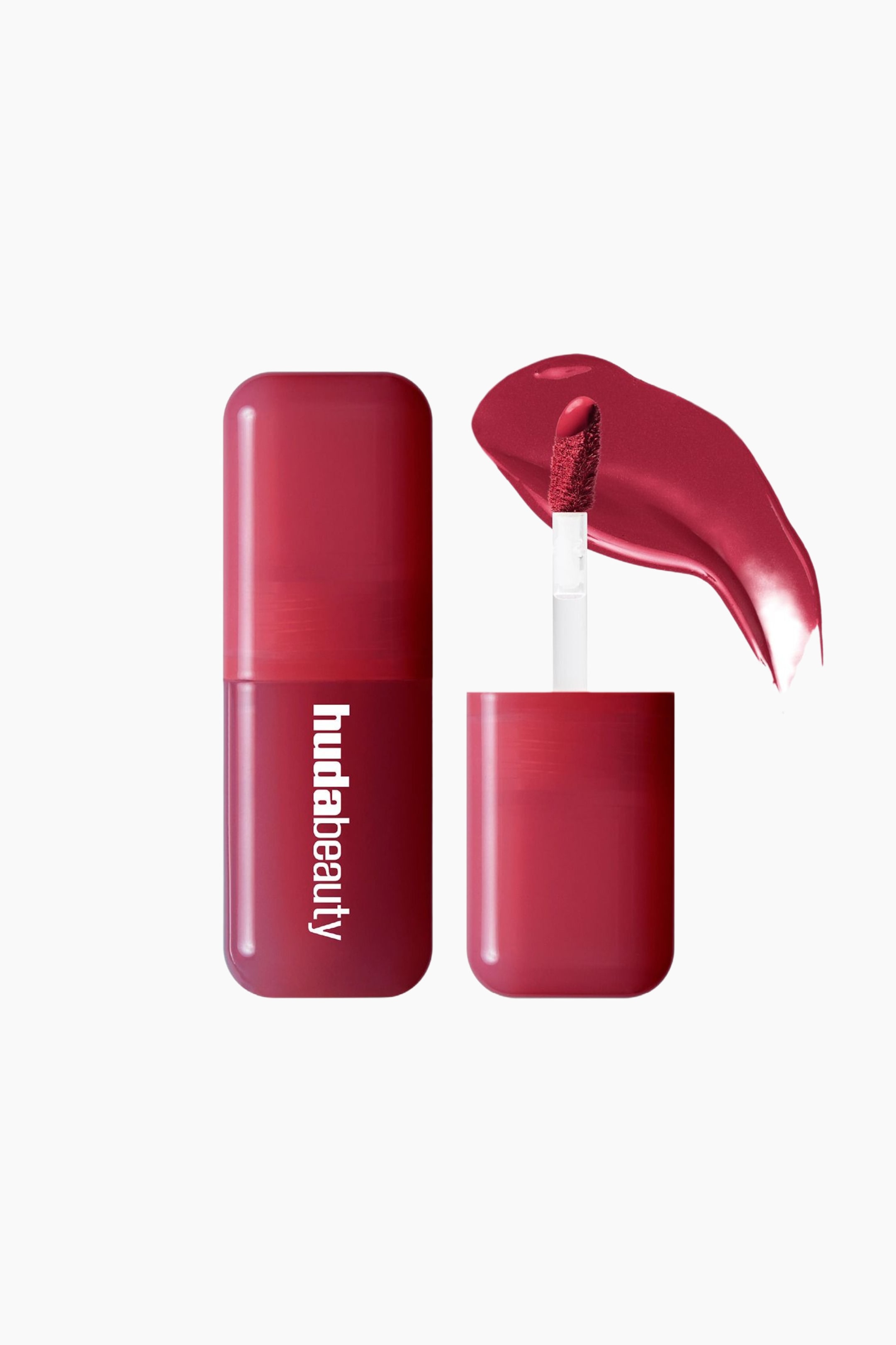 Visa större bild: En djupt rödrosa Huda Beauty flytande läppstiftsflaska står upprätt och visar "hudabeauty" vertikalt. En applikator med en flockad spets, täckt i den matchande vätskan, stiger upp ur den öppna flaskan, och en böjd provbit av vätskan syns till höger.