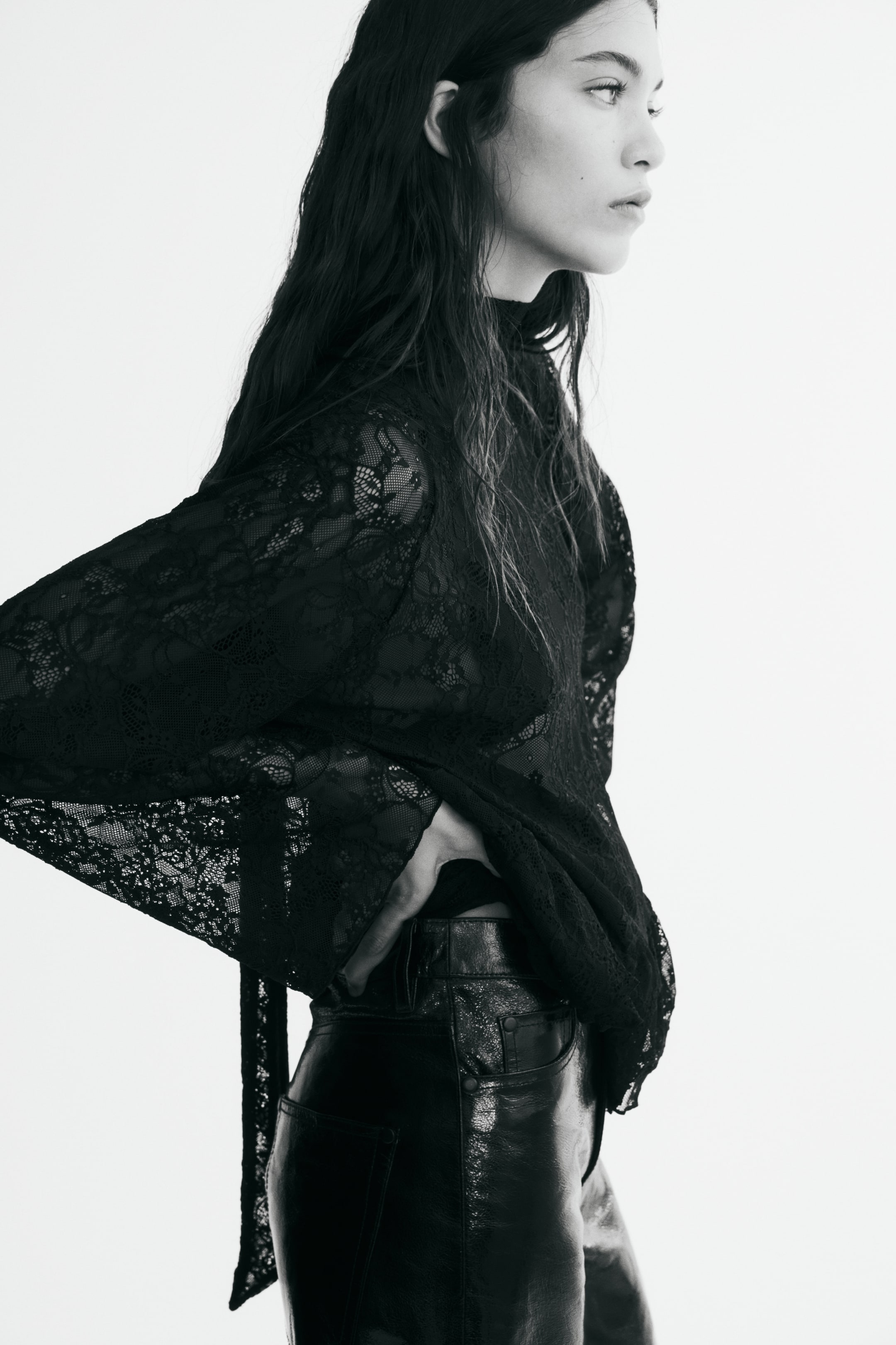 Grotere afbeelding bekijken: Een zwarte, semi-transparante kanten blouse met hoge hals en uitlopende mouwen wordt gedragen door het model. De top is gestyled met glanzende, getextureerde zwarte broek, waarbij het model één hand op de taille laat rusten.