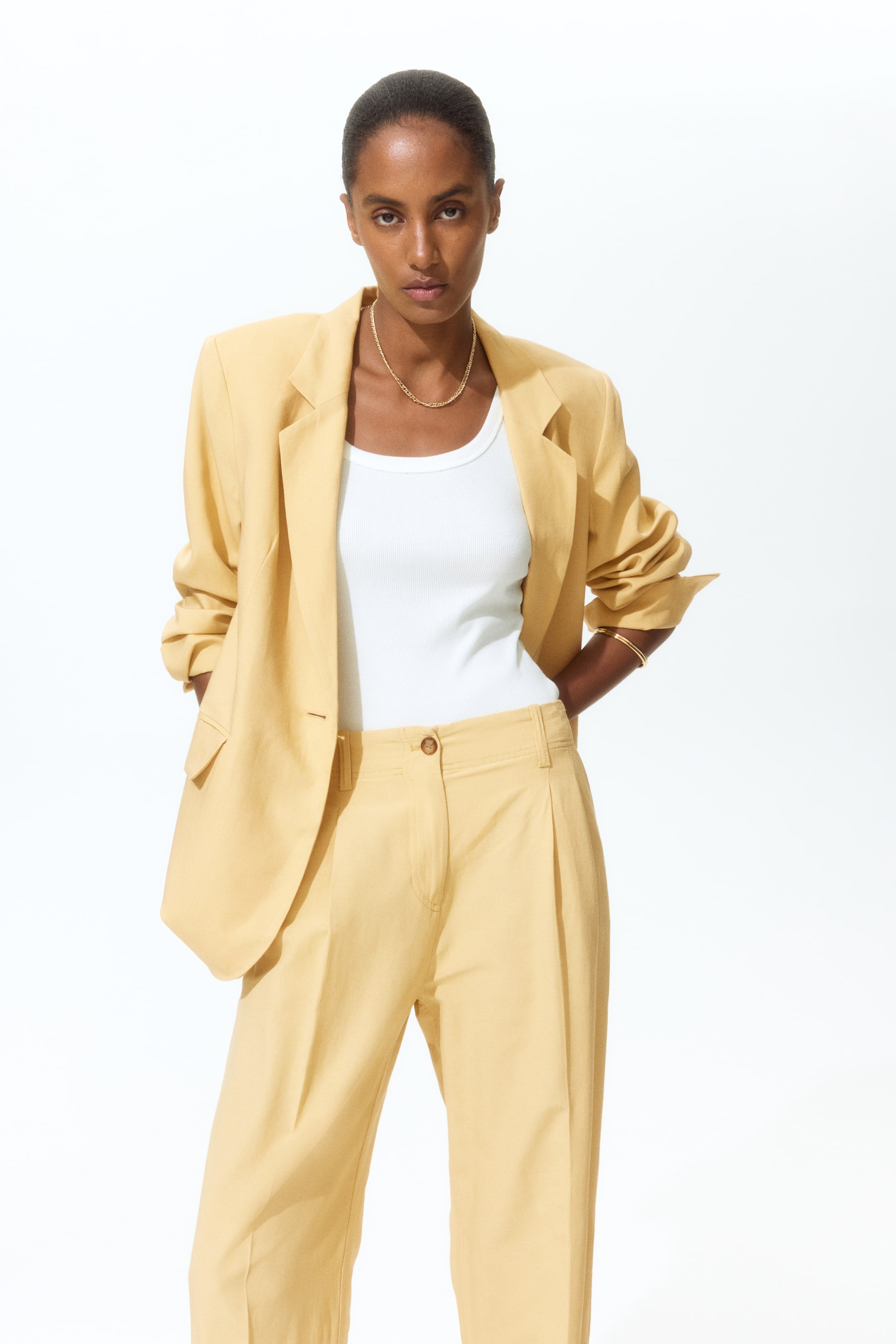 View larger image: A pale yellow single-breasted blazer with notch lapels and matching pleated trousers create a coordinated ensemble, facing forward. The blazer features a one-button closure and flap pockets, with its long sleeves rolled up. A white ribbed tank top is worn underneath, complemented by a gold chain necklace and gold bracelets to accessorize the look.