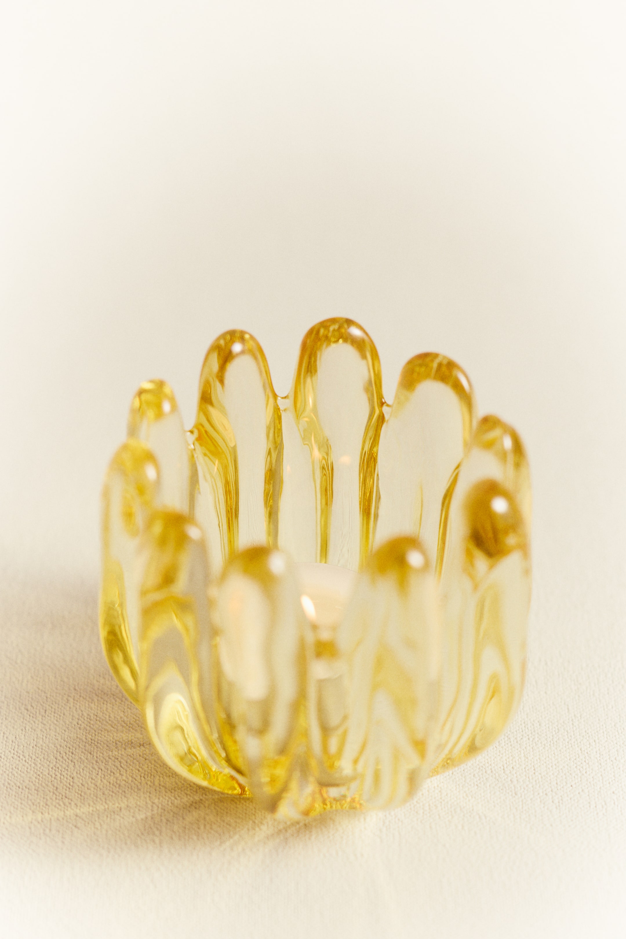 View larger image: "The glass tealight holder showcases a soothing amber tint and a design reminiscent of petals, featuring distinct, elevated, rounded glass segments that form its edge. Inside, a white tealight candle is clearly visible."