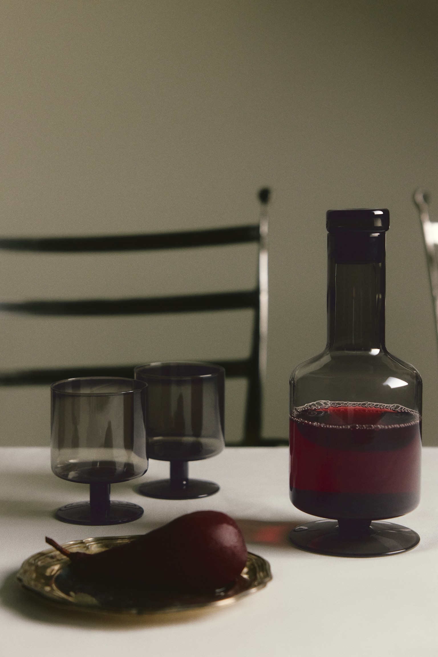 A footed wine decanter in smoky grey mouth-blown glass takes centre stage. It features a wide body with softly rounded edges, a narrow neck, and a dark glass stopper, resting on a dark, circular foot. - 1