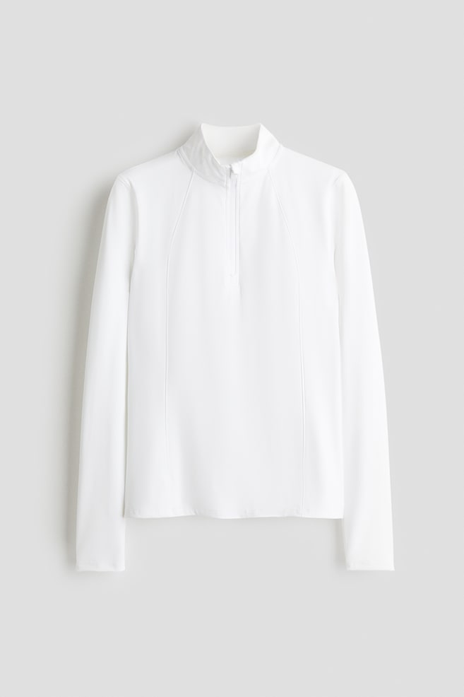 A white long-sleeved top is being presented from the front, featuring a standing collar and a quarter-zip front closure. Vertical seam lines are visible along the raglan sleeves and running down the front of the garment. - 1
