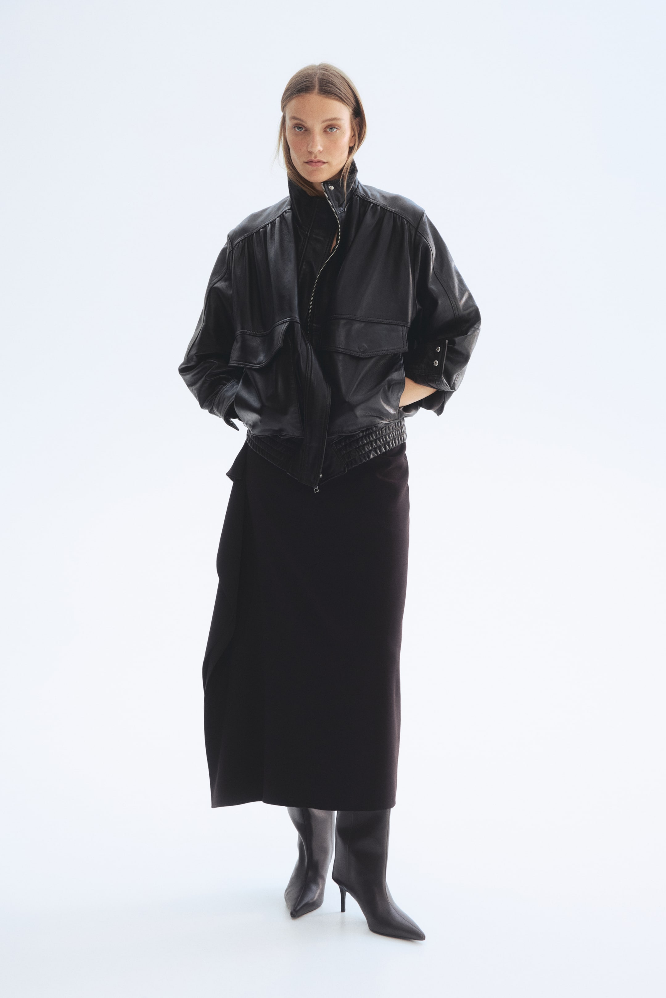 Büyük resmi görüntüle: Ön cepheden sunulan siyah deri ceket, yüksek bir dik yaka ve kısmen açık ön fermuar ile dikkat çekiyor. Ayrıca düşürülmüş omuzlar, düğme detaylı manşetlere sahip toplanmış kollar, göğüste iki büyük kapaklı cep ve beli sıkan lastikli bir etek ucu bulunuyor.
