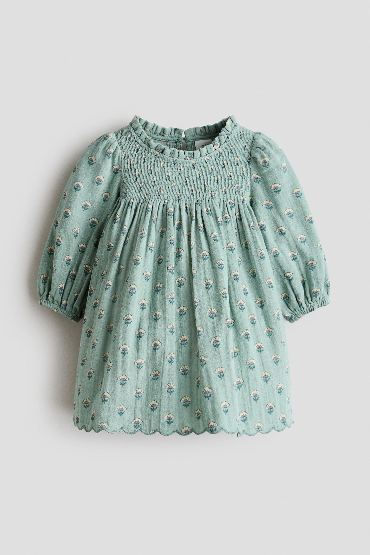 A front-facing baby dress is featuring a light green fabric with an all-over floral print. It is showcasing a ruffled high neck, a smocked bodice, long puffed sleeves with gathered cuffs, and a scalloped hemline. - 1