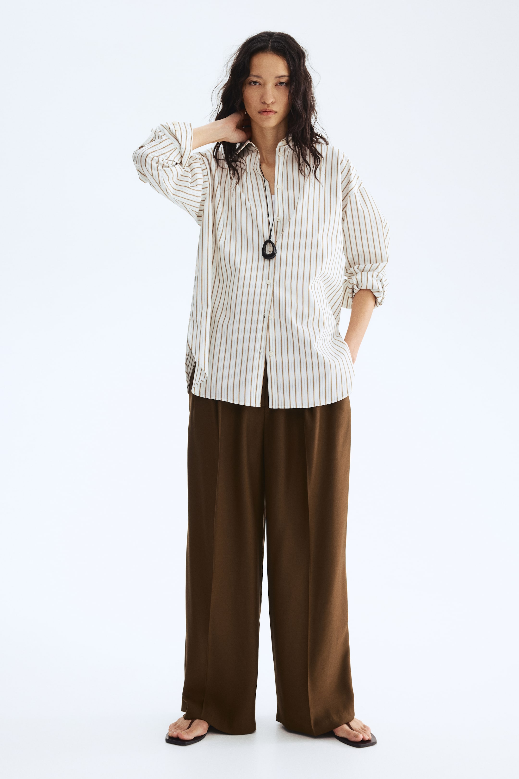View larger image: Dark brown wide-leg trousers are showcasing a laid-back, flowing silhouette, reaching the floor and partially concealing black thong sandals. They boast distinct vertical creases lining the front of each leg.