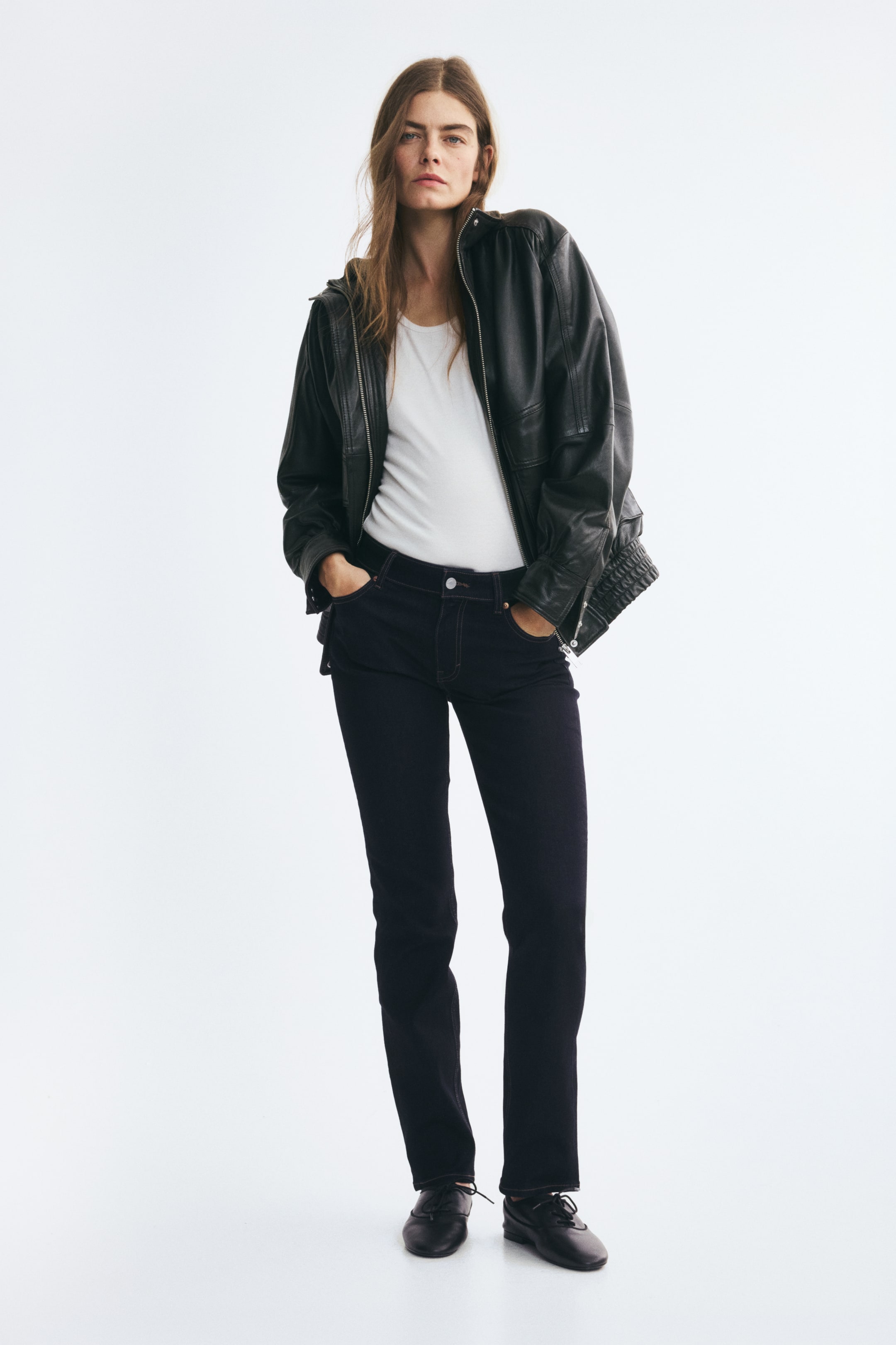 Größeres Bild ansehen: Dunkel gewaschene Denim-Jeans mit Kontraststeppnähten und normaler Bundhöhe werden vom Model getragen, das mit den Händen in den Vordertaschen steht. Die Slim-Fit-Jeans sind mit einem in den Bund gesteckten weißen T-Shirt, einer offenen schwarzen Lederjacke und schwarzen Schnürschuhen kombiniert.