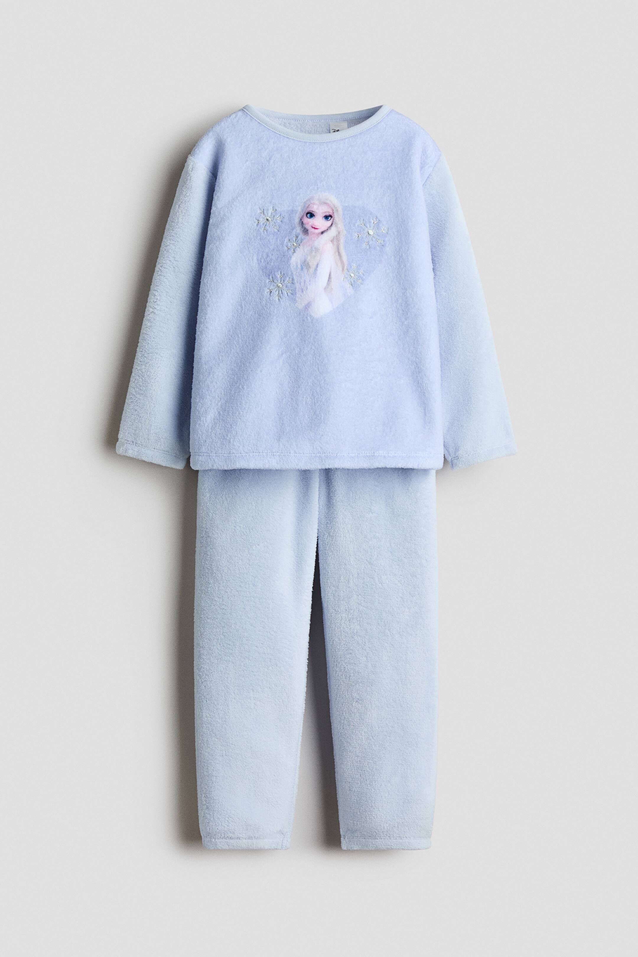 View larger image: A light blue velour pyjama set is shown from the front, featuring a long-sleeved top with a round neckline and a central graphic print of Elsa from Frozen. The matching trousers are displayed below, showing a soft, tapered fit with an elasticated waistband.