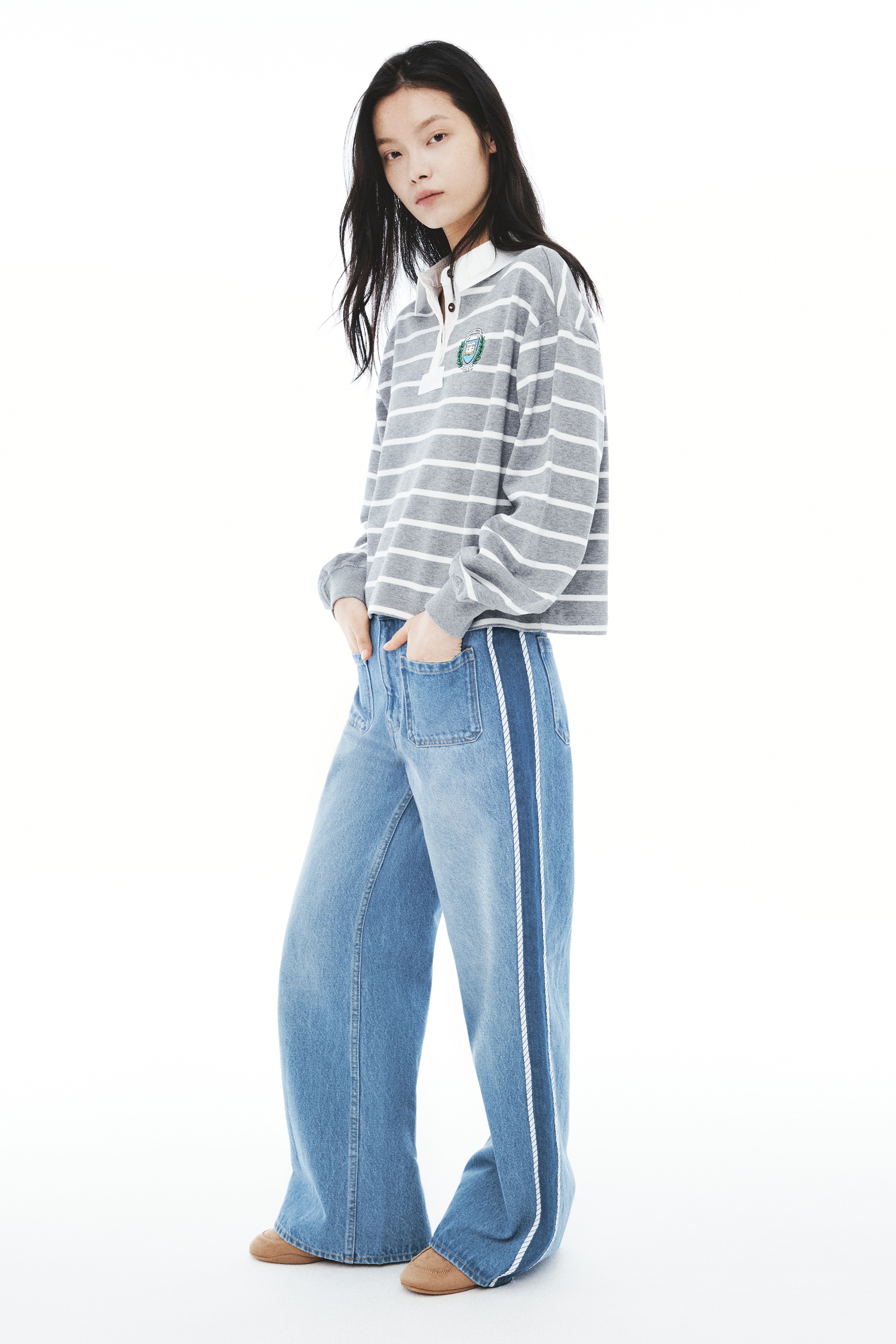 A pair of wide-leg blue jeans is facing front, featuring a medium wash with faded detailing down the centre of the legs and two front patch pockets below the waistband. White rope-like piping runs vertically down each side seam, separating the main body of the jeans from slightly darker blue denim side panels.
