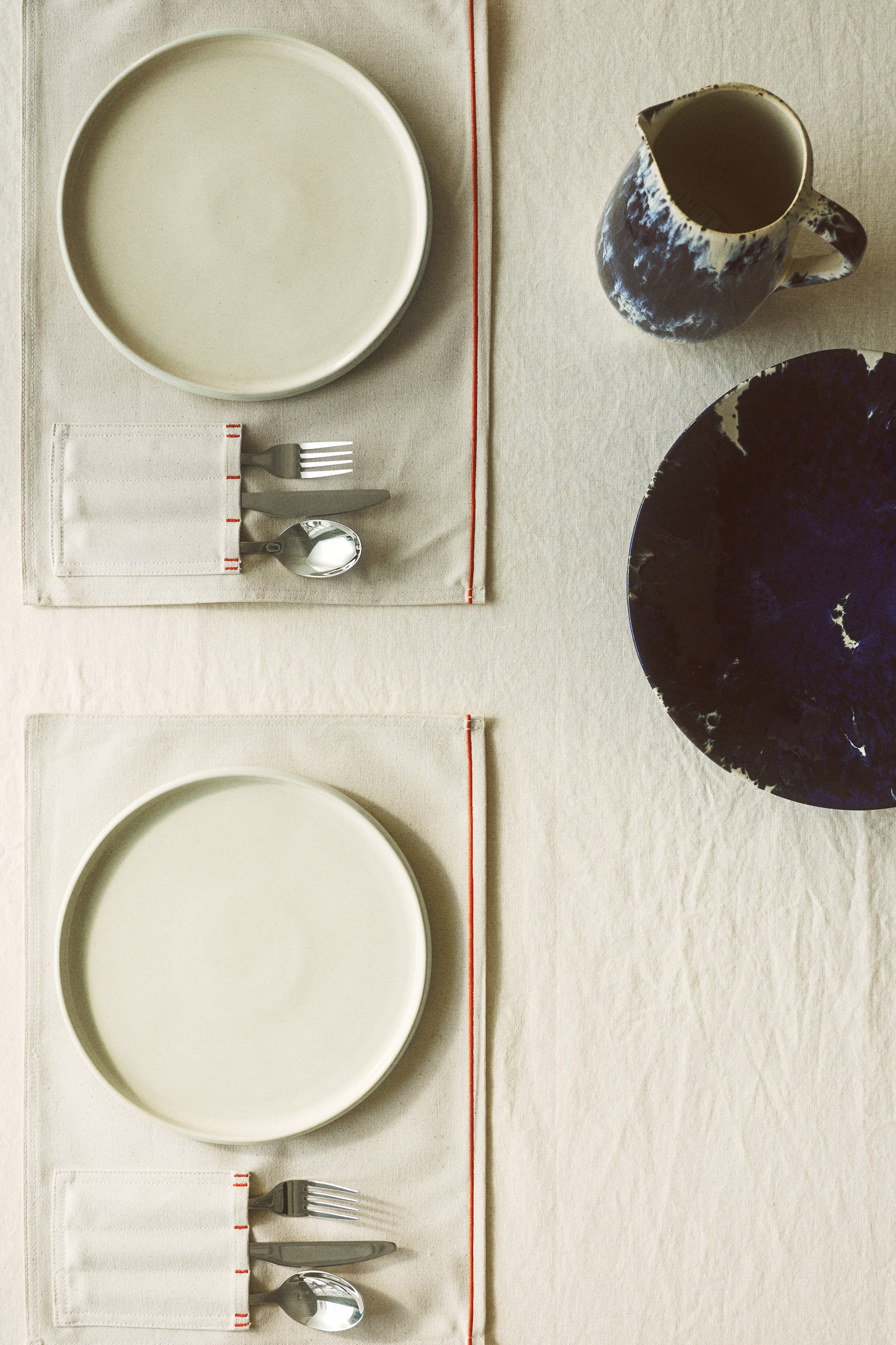 Agrandir l'image: Deux ensembles de couverts individuels présentent des assiettes rondes de couleur crème sur des sets de table en tissu léger, chaque set de table arborant une rayure rouge verticale et une poche contenant une fourchette, un couteau et une cuillère avec des manches sombres. Une carafe en céramique à motifs bleus et blancs et un bol profond sont également visibles sur la table, le tout vu de dessus.
