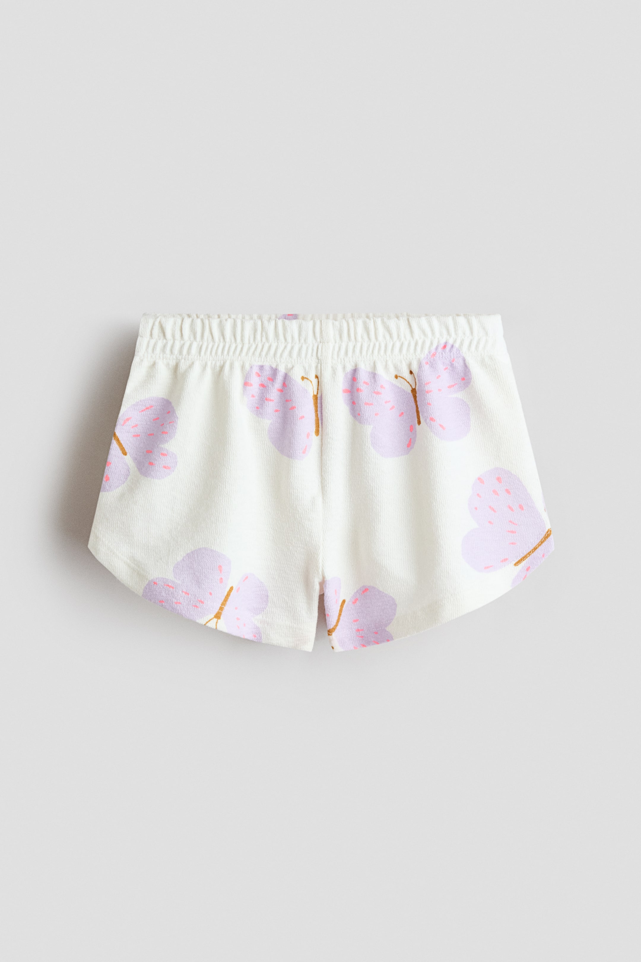 Agrandir l'image: Short en jersey de coton texturé avec taille élastiquée présenté de face. Le tissu blanc est orné de papillons violets clairs, agrémentés de taches roses et de corps fins marron.