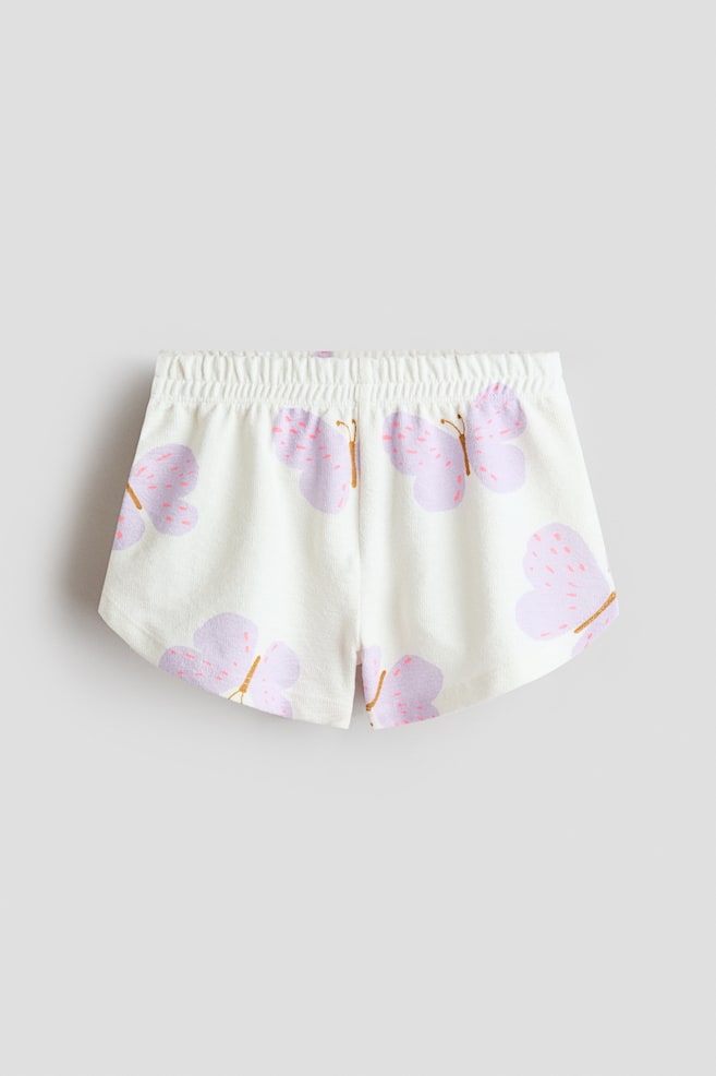 Shorts in textured cotton jersey with an elasticated waist are presenting a front view. The white fabric is patterned with light purple butterflies, which are accented with pink speckles and thin brown bodies. - 1
