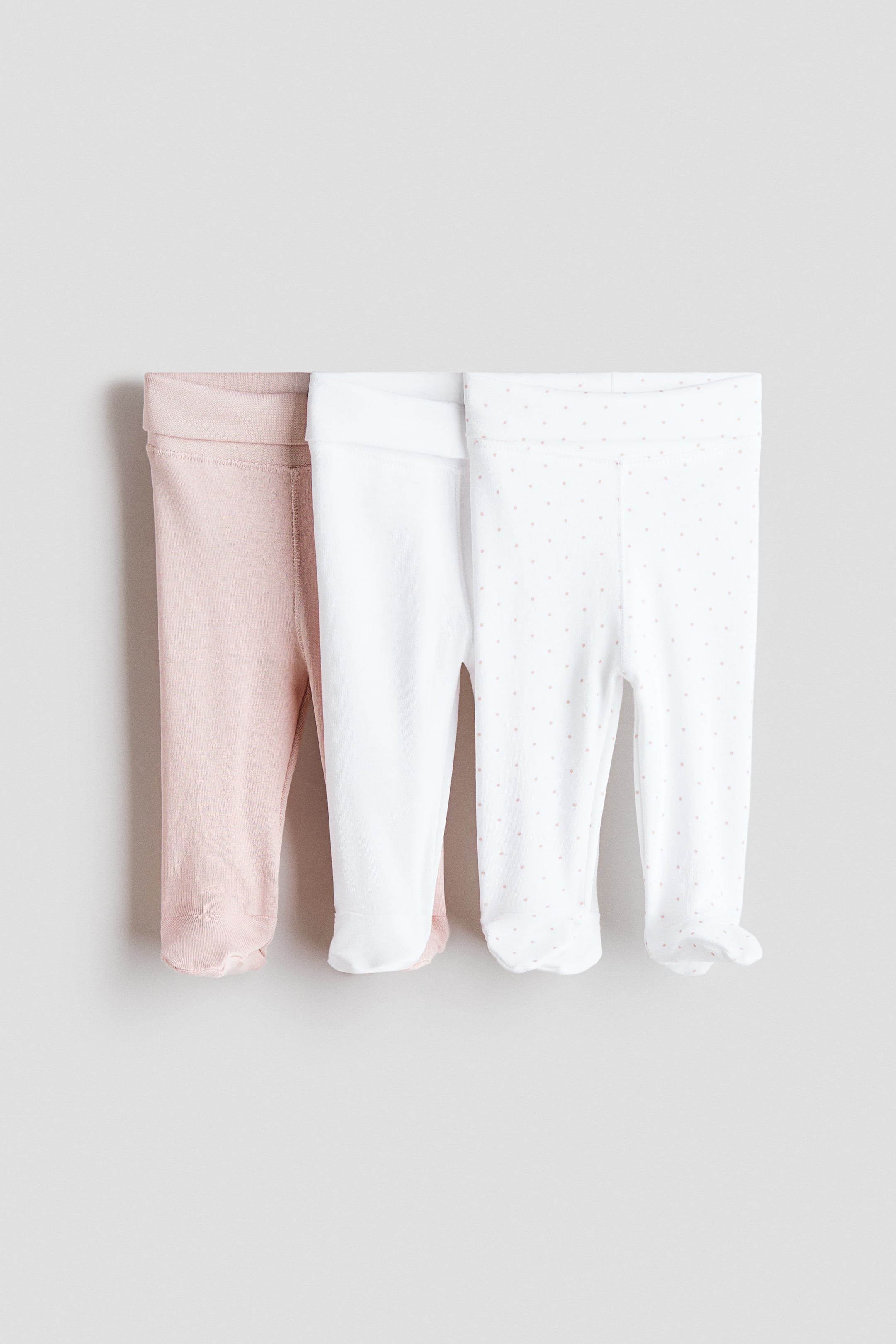 View larger image: Three pairs of ultra-soft cotton jersey leggings are displayed, viewed from the front, each showcasing a broad, foldover waist panel and full feet with elasticated heels. The set comprises a light pink pair, a white pair, and a white pair adorned with small pink polka dots.