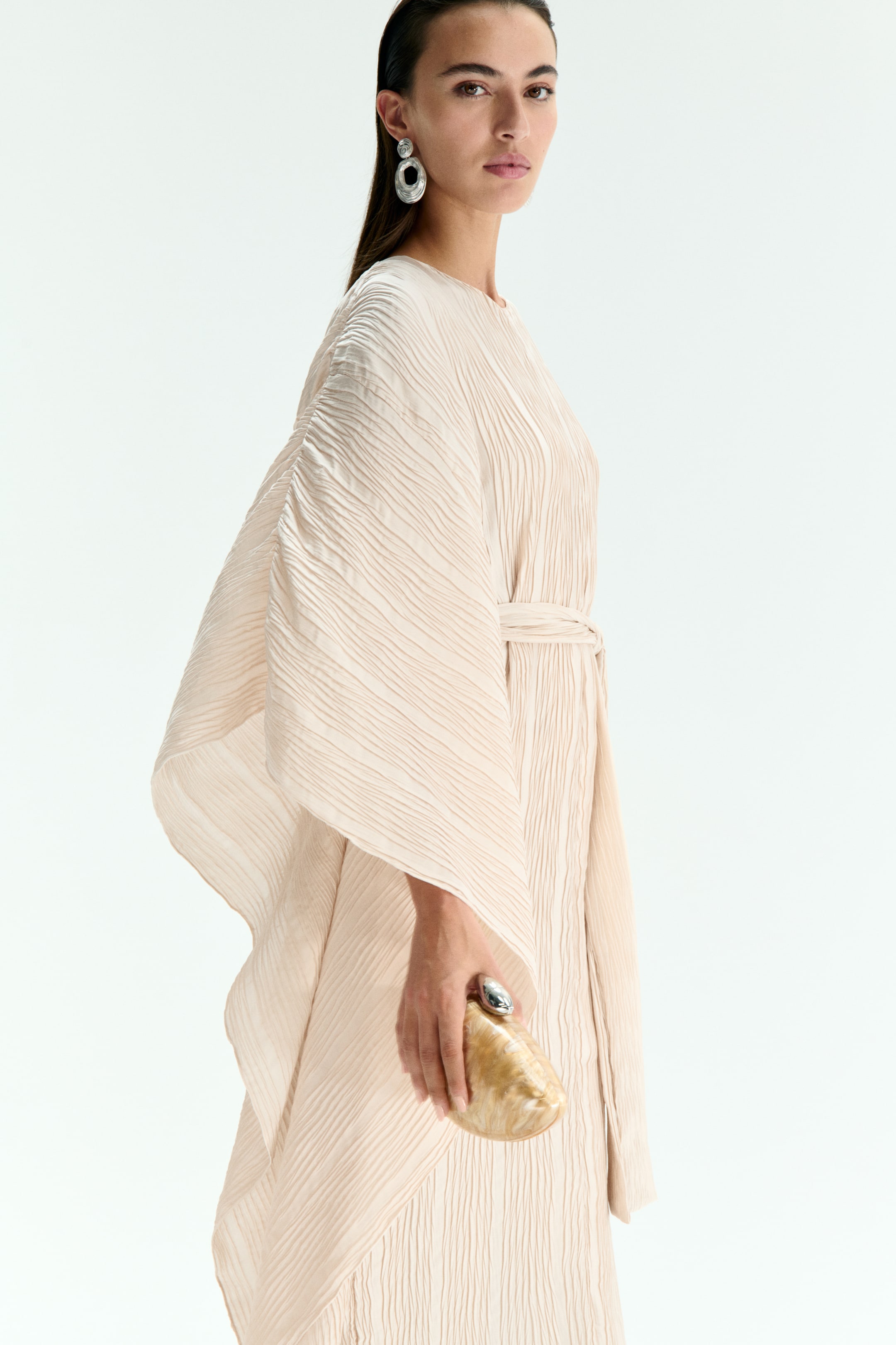 View larger image: A light beige pleated kaftan dress, featuring wide sleeves and a matching self-tie belt cinching the waist, is being worn by the model. The model is holding a small, pearlescent brown clutch bag.