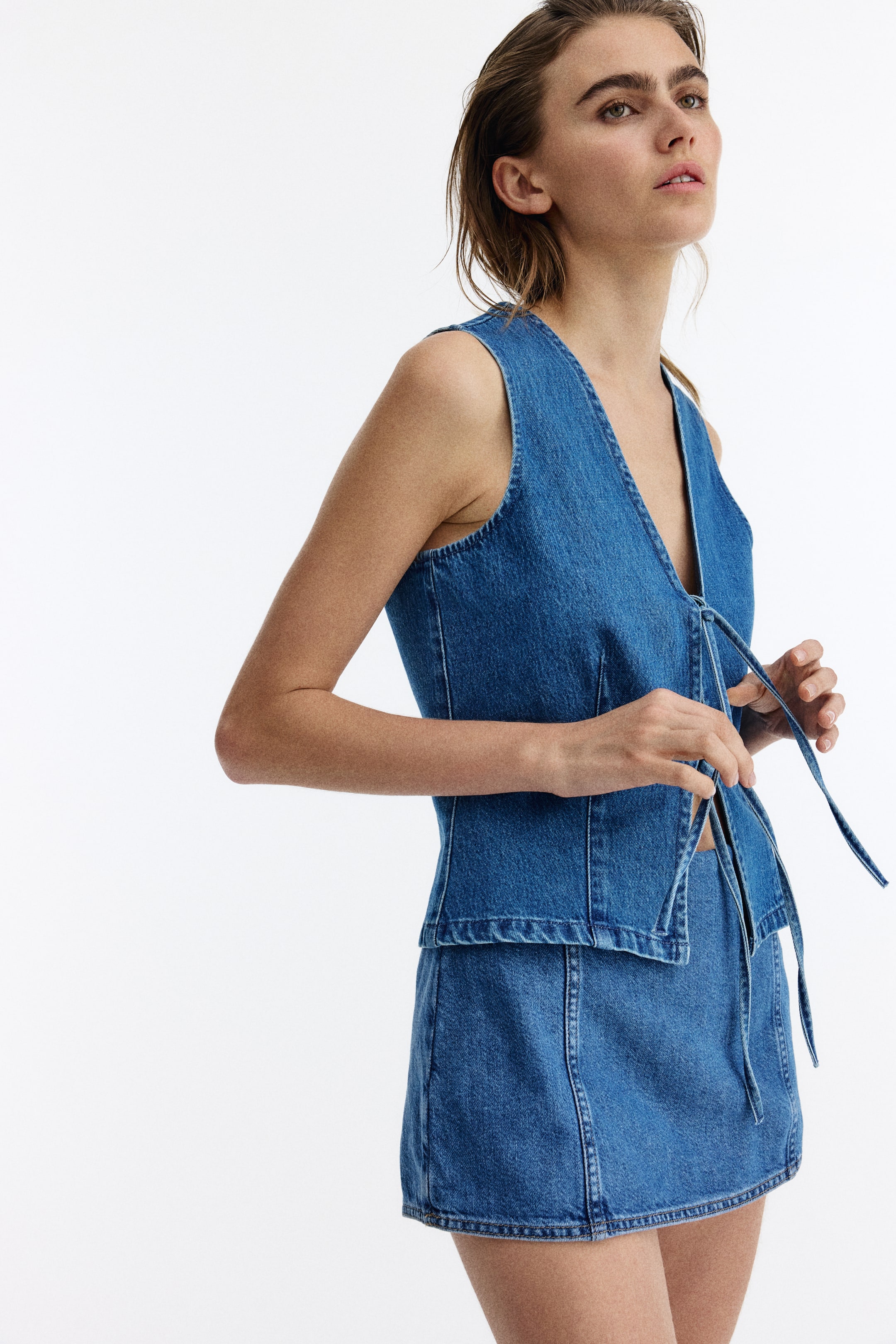 View larger image: A tailored denim cotton waistcoat features a V-neckline and two sets of slim ties at the front, angled to the left to emphasize its fitted design.