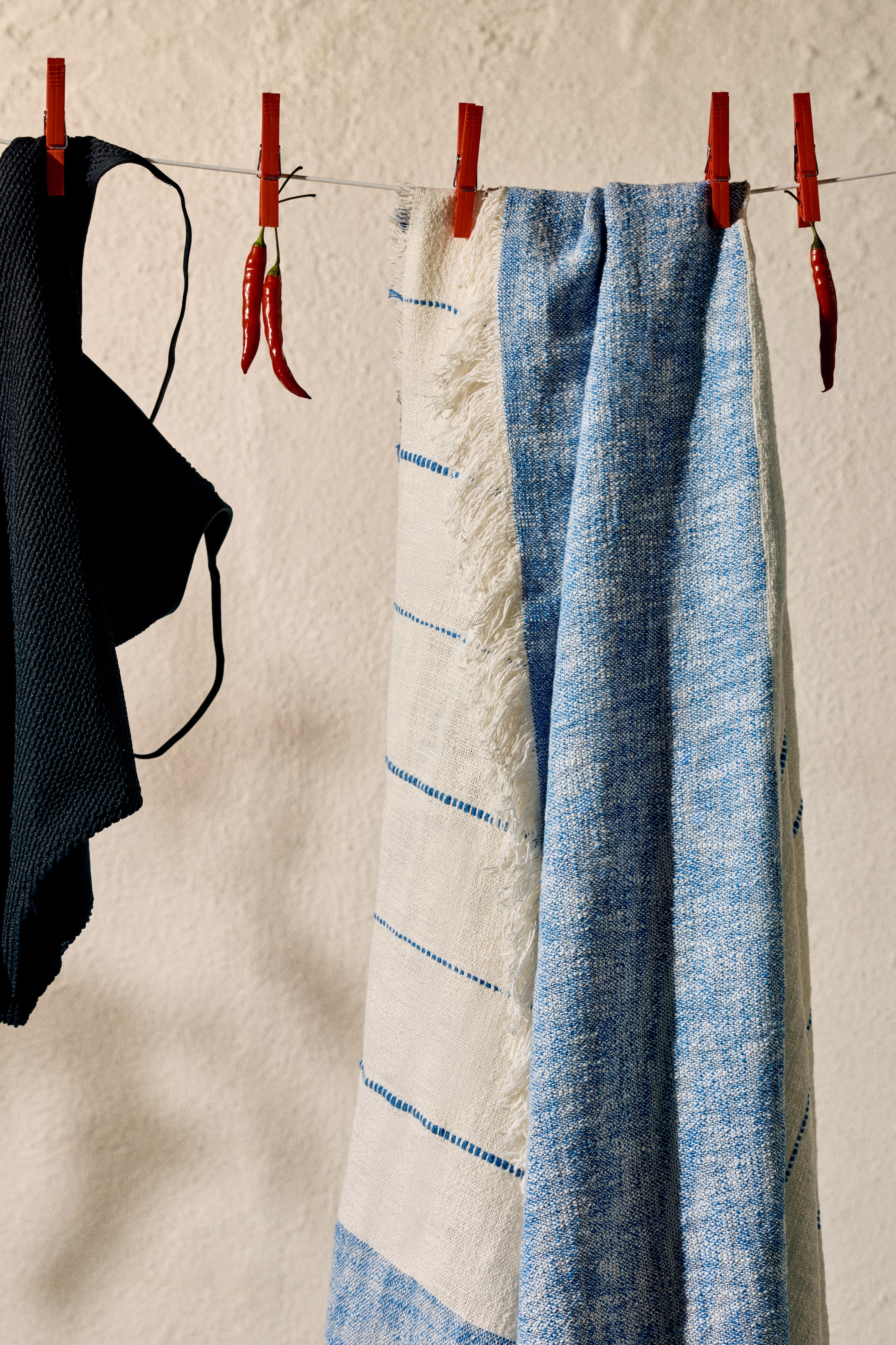 View larger image: A dark, textured fabric piece with a visible strap is hanging. Two woven textiles are draped beside it; one features white with blue dashed stripes and fringed edges, while the other showcases a mottled blue and white weave.