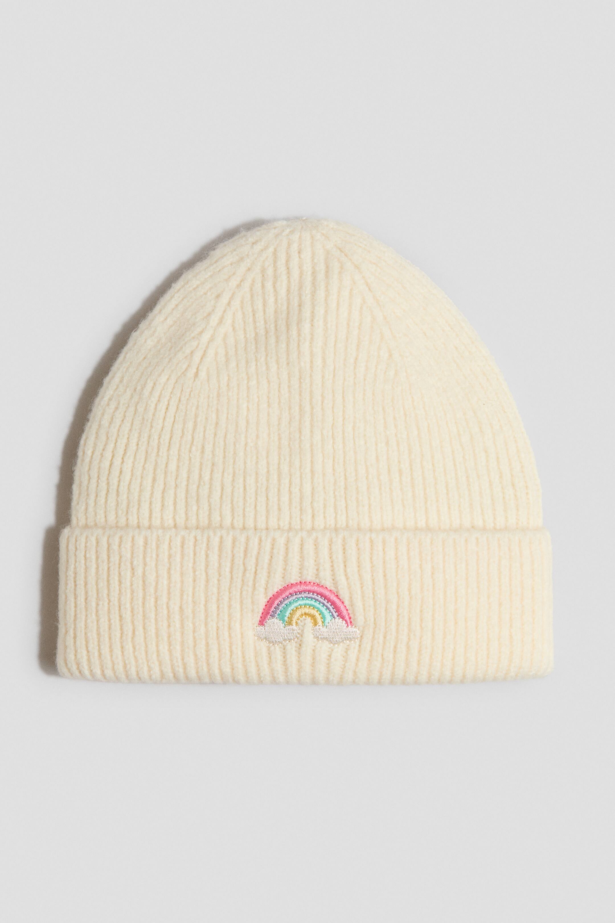 Ver imagen más grande: Un gorro de punto color crema aparece de frente, con textura acanalada y vuelta en el borde. En la parte frontal de la vuelta lleva bordado un diseño de arcoíris colorido con pequeñas nubes blancas.
