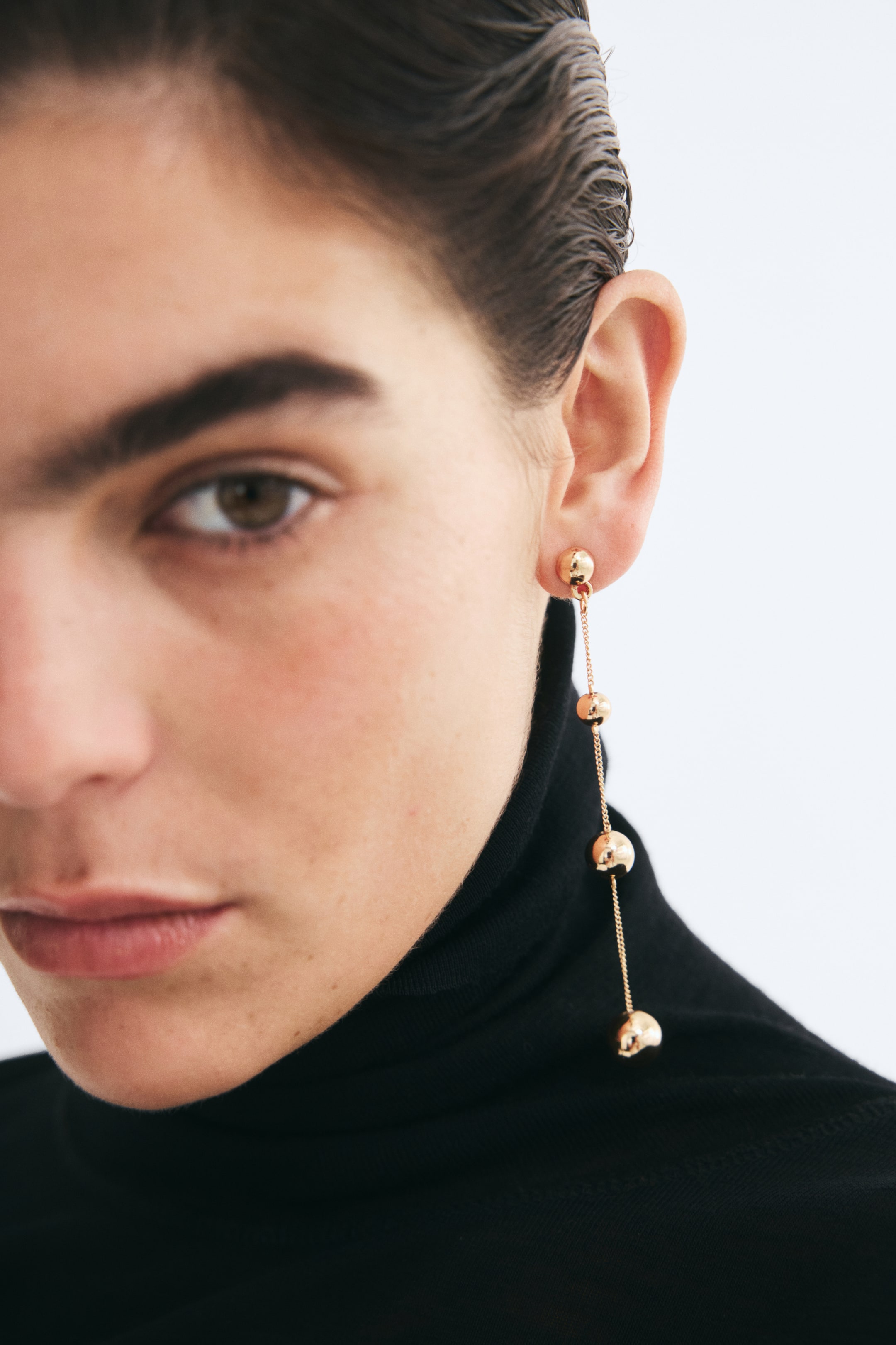 View larger image: A long metal chain earring featuring evenly spaced gold-toned plastic beads that increase in size toward the bottom is worn on the model’s ear. The earring hangs down alongside a black turtleneck top.