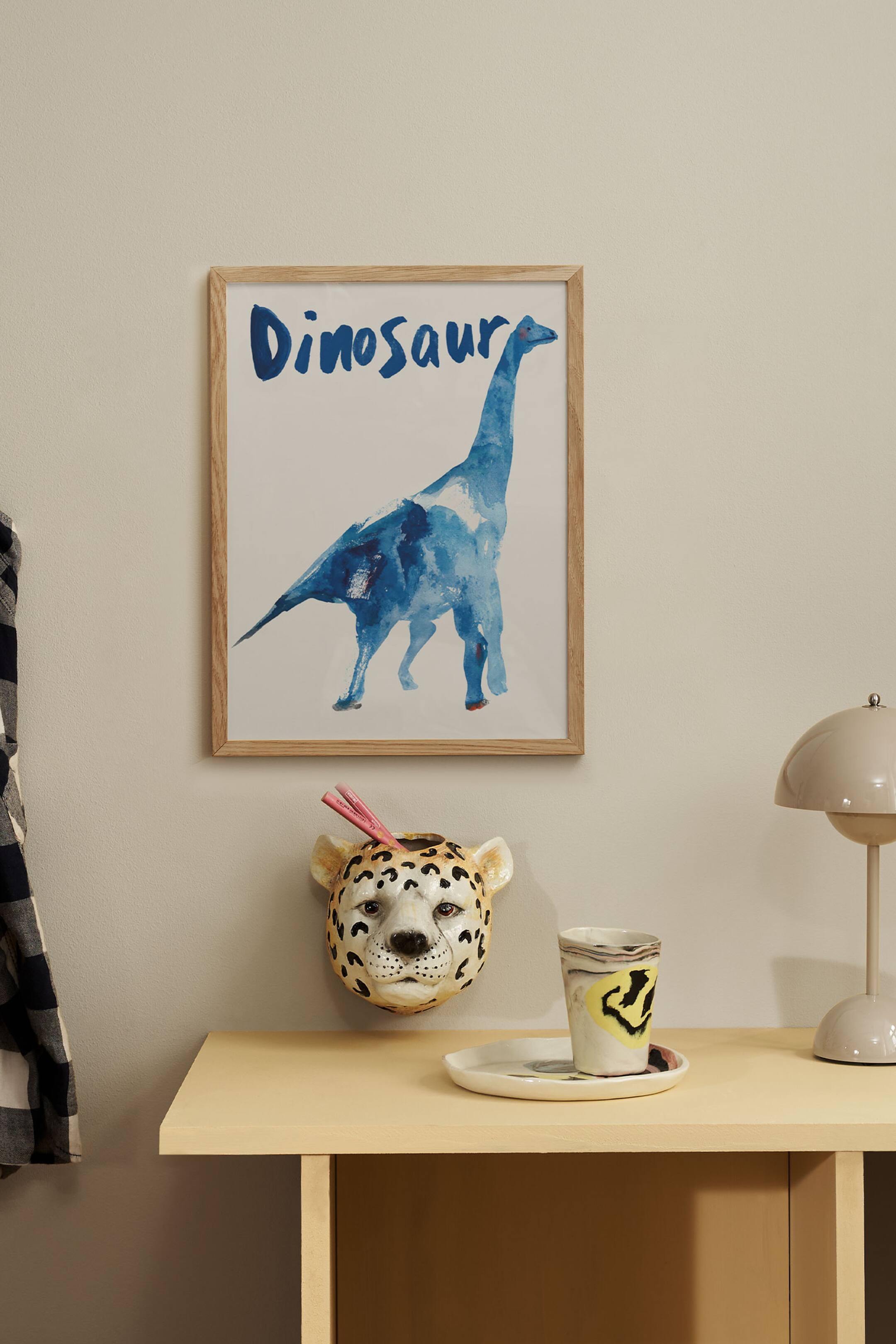Grotere afbeelding bekijken: Een rechthoekige ingelijste kunstprint met een aquarelillustratie van een blauwe dinosaurus met lange nek op een effen witte achtergrond. Het woord "Dinosaur" staat bovenaan in blauw, met de hand geschilderde letters, en de print is omlijst met een lichtgekleurde houten lijst.