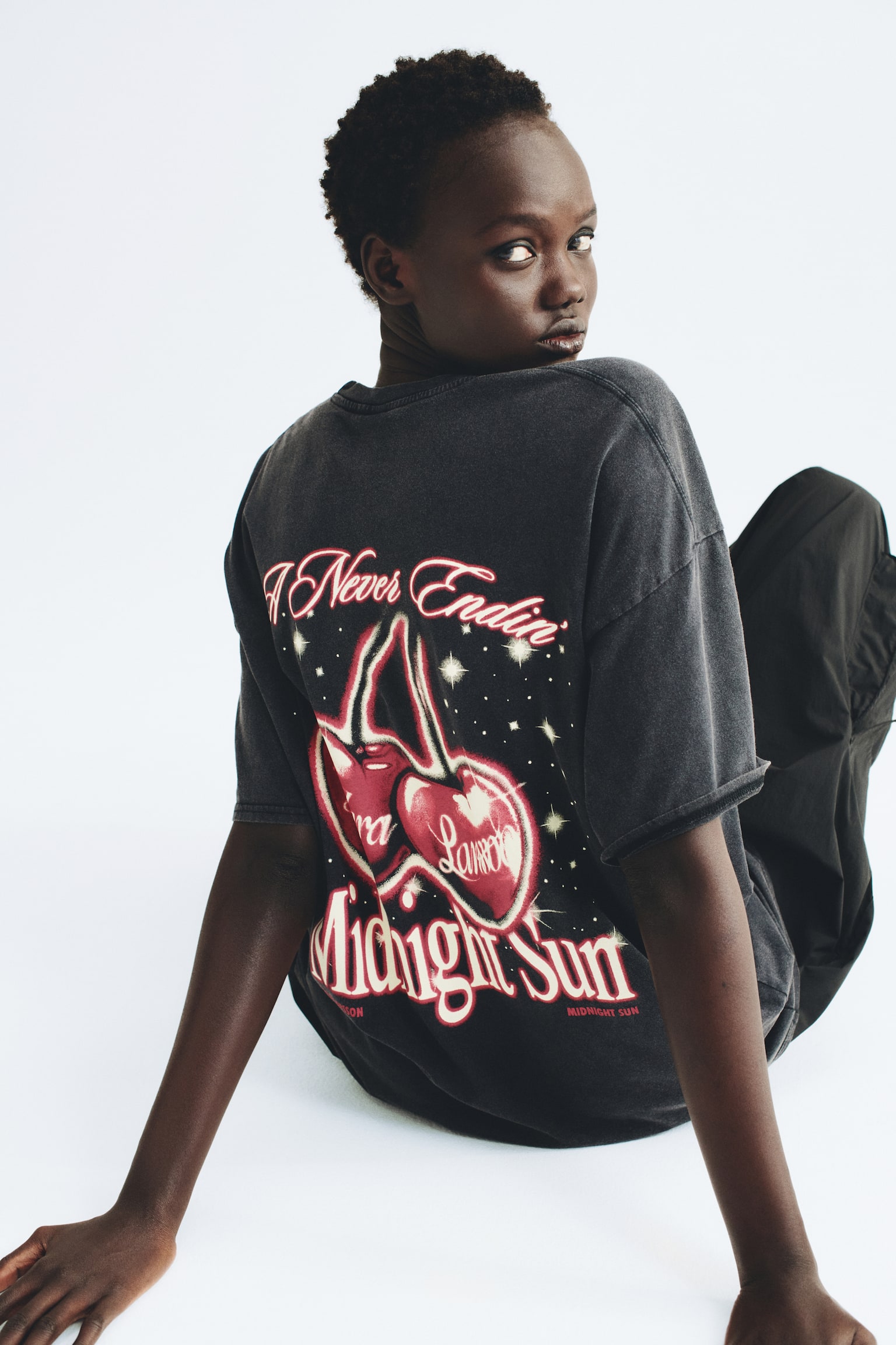 En oversized, tvÃ¤ttad svart t-shirt med nedhasade axlar bÃ¤rs av modellen, som sitter och tittar bak Ã¶ver axeln. T-shirten har ett stort grafiskt tryck pÃ¥ ryggen som fÃ¶restÃ¤ller rÃ¶da kÃ¶rsbÃ¤r och stiliserad text. - 1