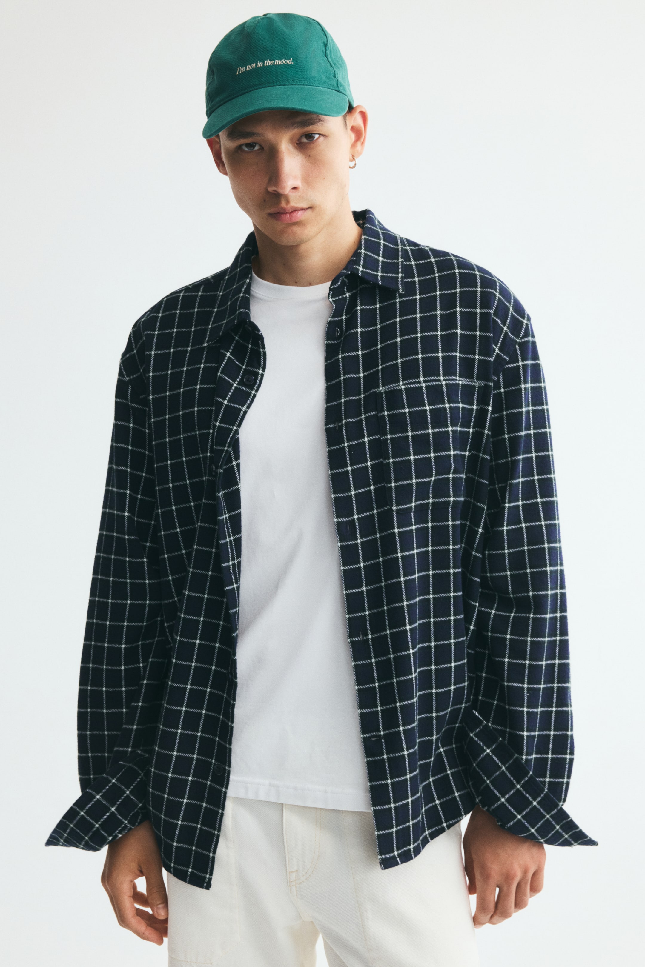 Se større billede: En flannelshirt med navyblåt og hvidt gittermønster bæres åbent af modellen over en hvid T-shirt. Den afslappede skjorte har klassisk krave, sænkede skuldre, en enkelt påsyet brystlomme og lange ærmer med manchetter uden knapper.