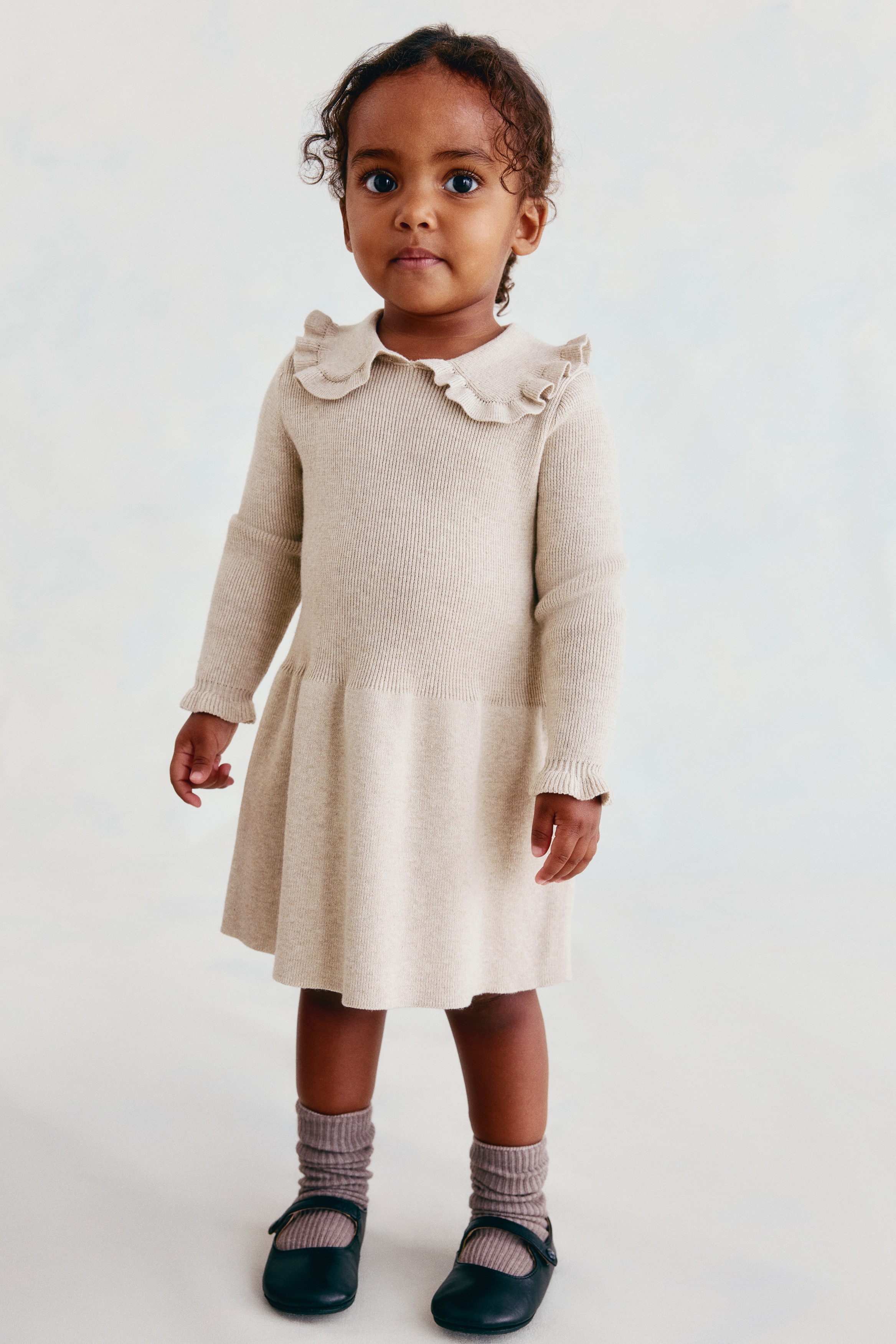 A beige knit dress is featuring a dense rib knit bodice, a rounded frill-trimmed collar, and long sleeves with frilled cuffs. The dress is also showcasing a flared skirt and an opening visible at the back of the neck.