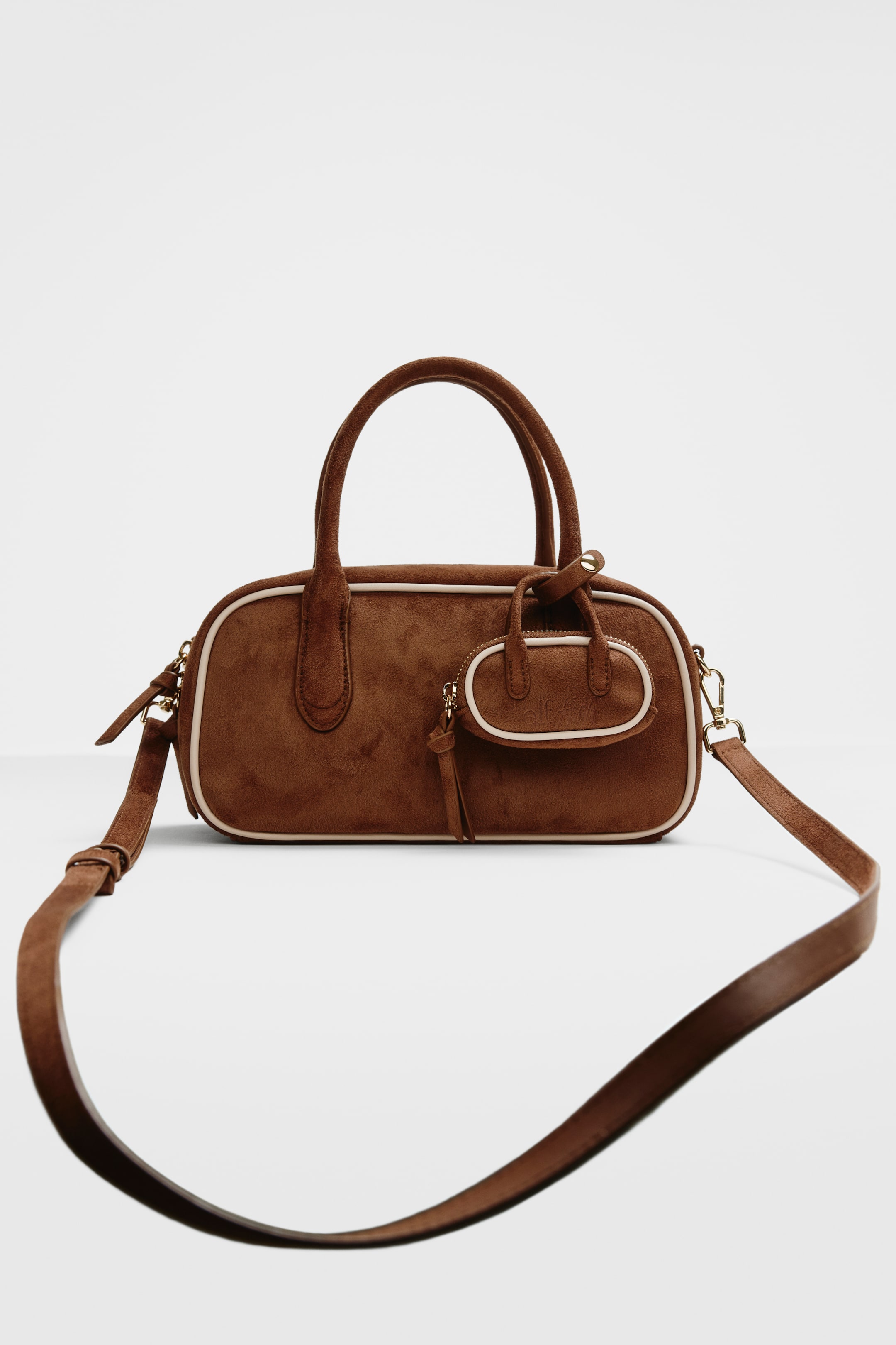 View larger image: A brown, suede-like duffle bag faces forward, showcasing an elongated, rounded body with white piping along its edges and two top handles. A small, oval-shaped pouch with a zipper and an "elf" logo is attached to the front, with a matching shoulder strap extending downward from the sides.