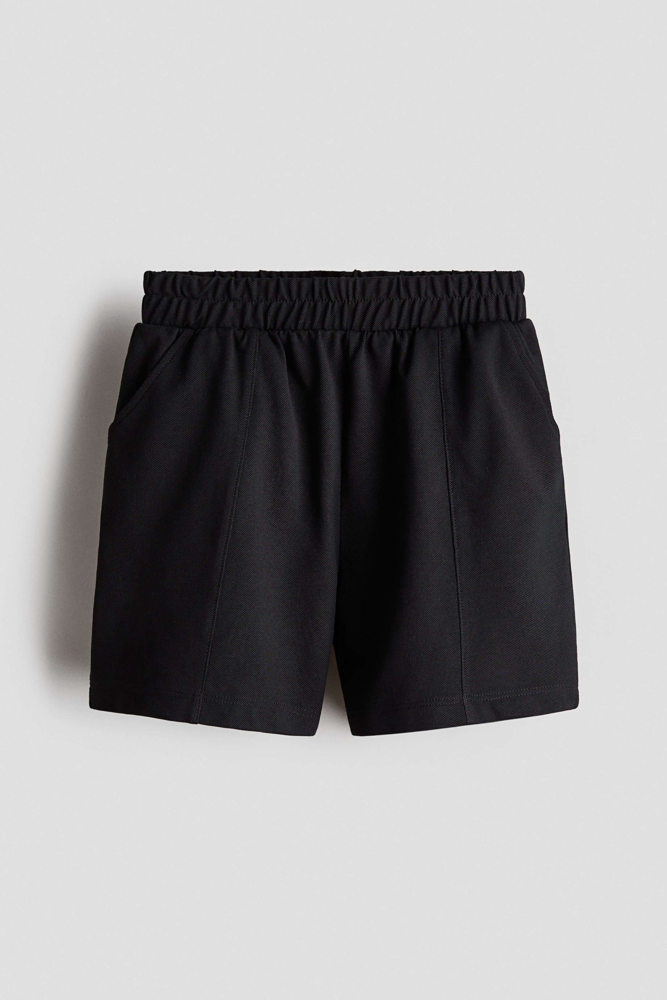 Xem ảnh lớn hơn: Quần short màu đen được chụp ở góc nhìn chính diện, nổi bật với cạp thun bản rộng và túi chéo hai bên. Thiết kế có cấu trúc với các đường may dọc chạy xuống phía trước mỗi ống quần.