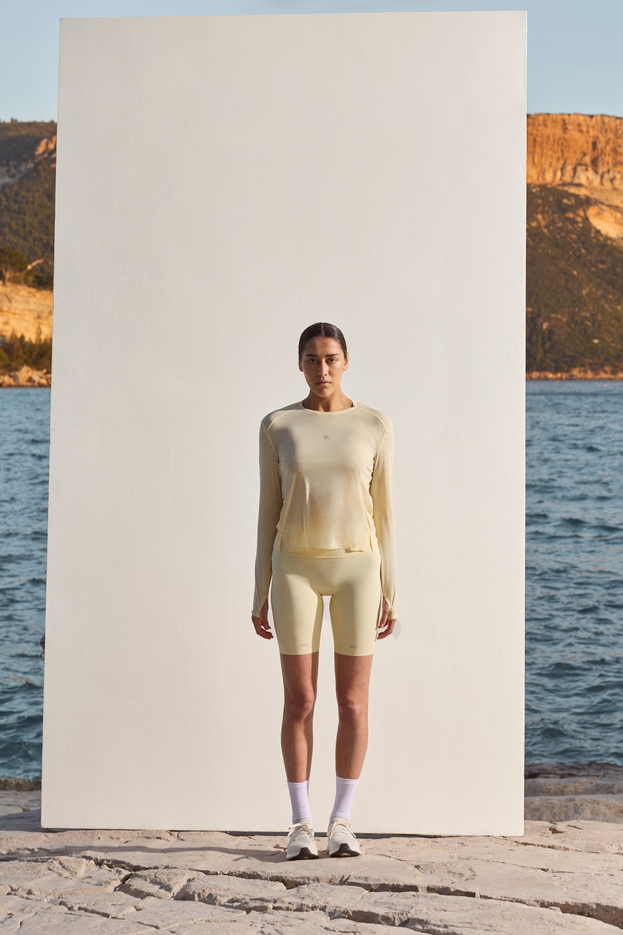 View larger image: A pale yellow long-sleeved top and matching cycling shorts are showcased from the front. The top features a crew neck, raglan sleeves with thumbholes, and a small chest logo, while the high-waisted shorts extend to mid-thigh with subtle branding on the thighs.
