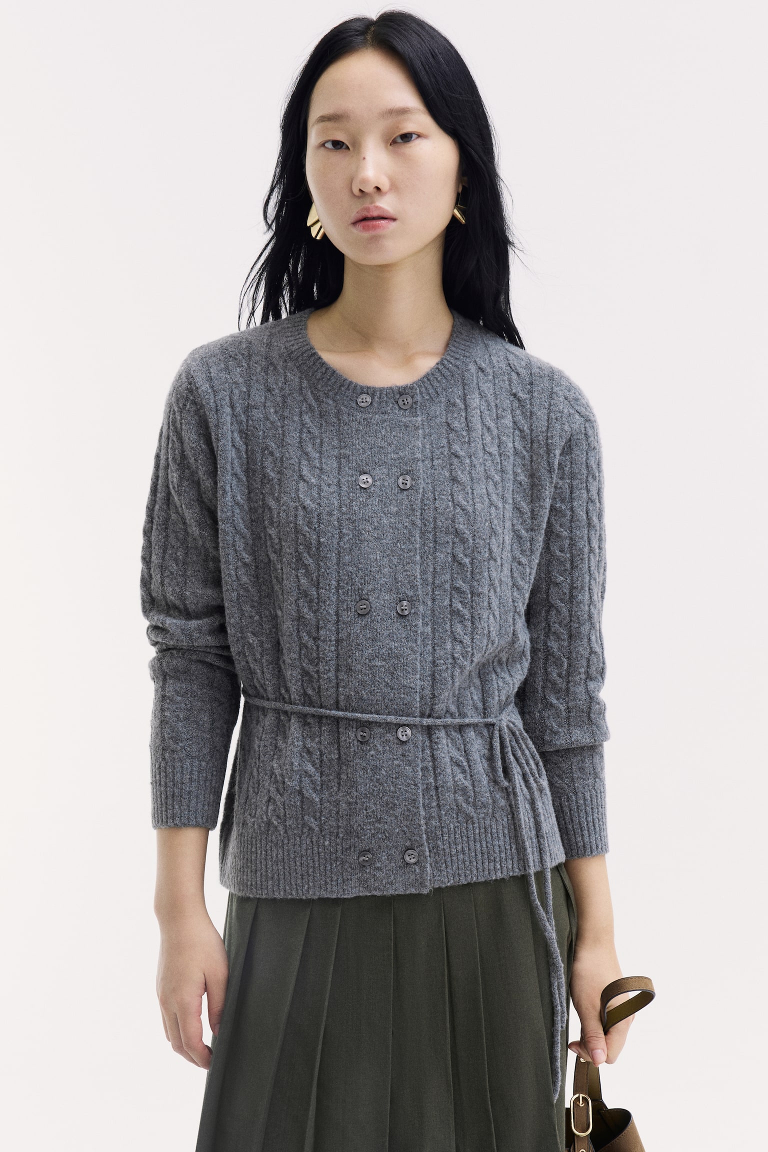 The model is wearing a gray, long-sleeved cable-knit cardigan featuring a round ribbed neckline, double-breasted front buttons, and a narrow tie belt at the waist. It is styled with a dark green pleated skirt, and the model is holding a brown bag strap. - 1