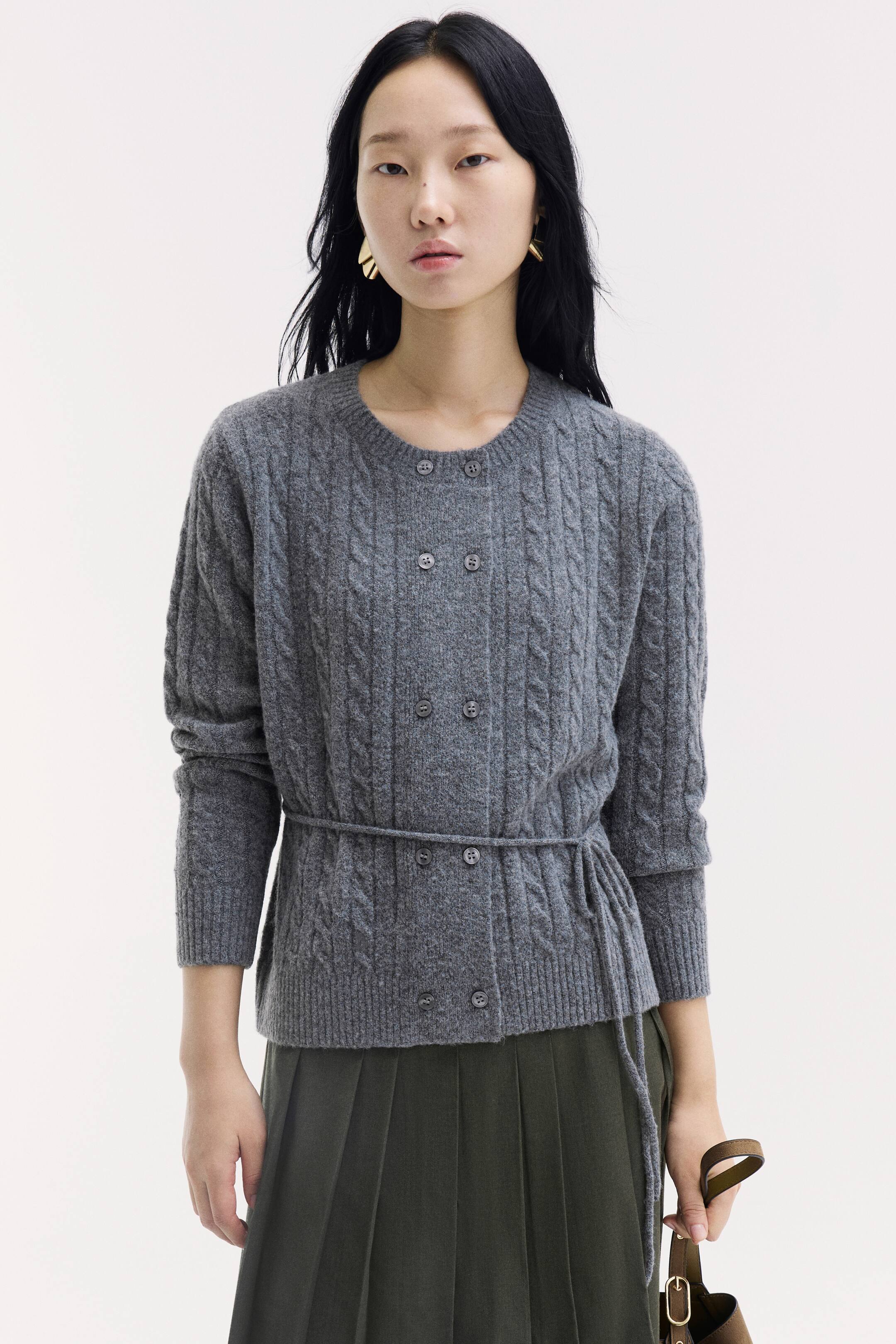 View larger image: The model is wearing a gray, long-sleeved cable-knit cardigan featuring a round ribbed neckline, double-breasted front buttons, and a narrow tie belt at the waist. It is styled with a dark green pleated skirt, and the model is holding a brown bag strap.