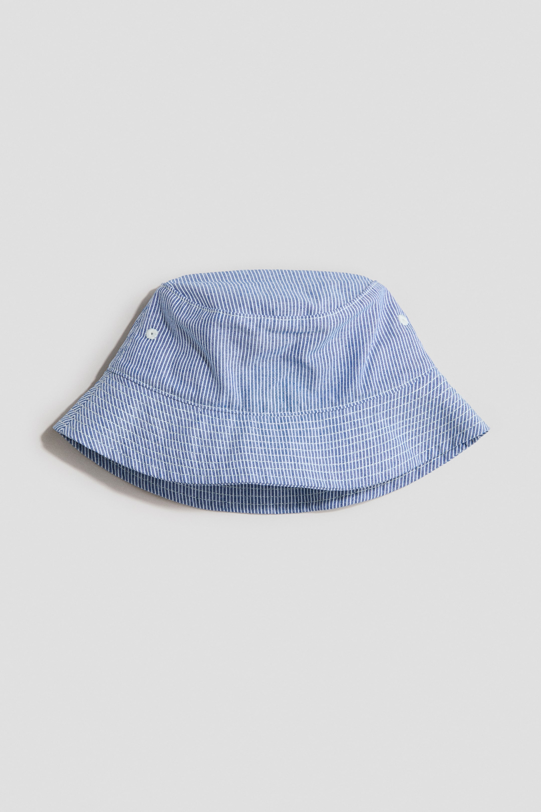 Zobrazit větší obrázek: Klobouk typu "bucket hat" je zobrazen z předního pohledu, vyrobený z modro-bílé pruhované bavlněné tkaniny s vertikálními pruhy na koruně a horizontálními pruhy na krempě. Viditelné šití zdobí krempu a na koruně jsou vidět dva malé bílé průduchy.