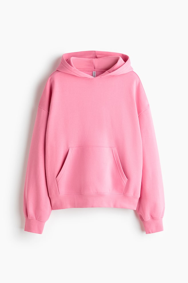 A pink fleece hoodie is displayed in a front-facing view, showing a jersey-lined hood, dropped shoulders, and long sleeves. The garment features a central kangaroo pocket with ribbed detailing at the cuffs and hem. - 1