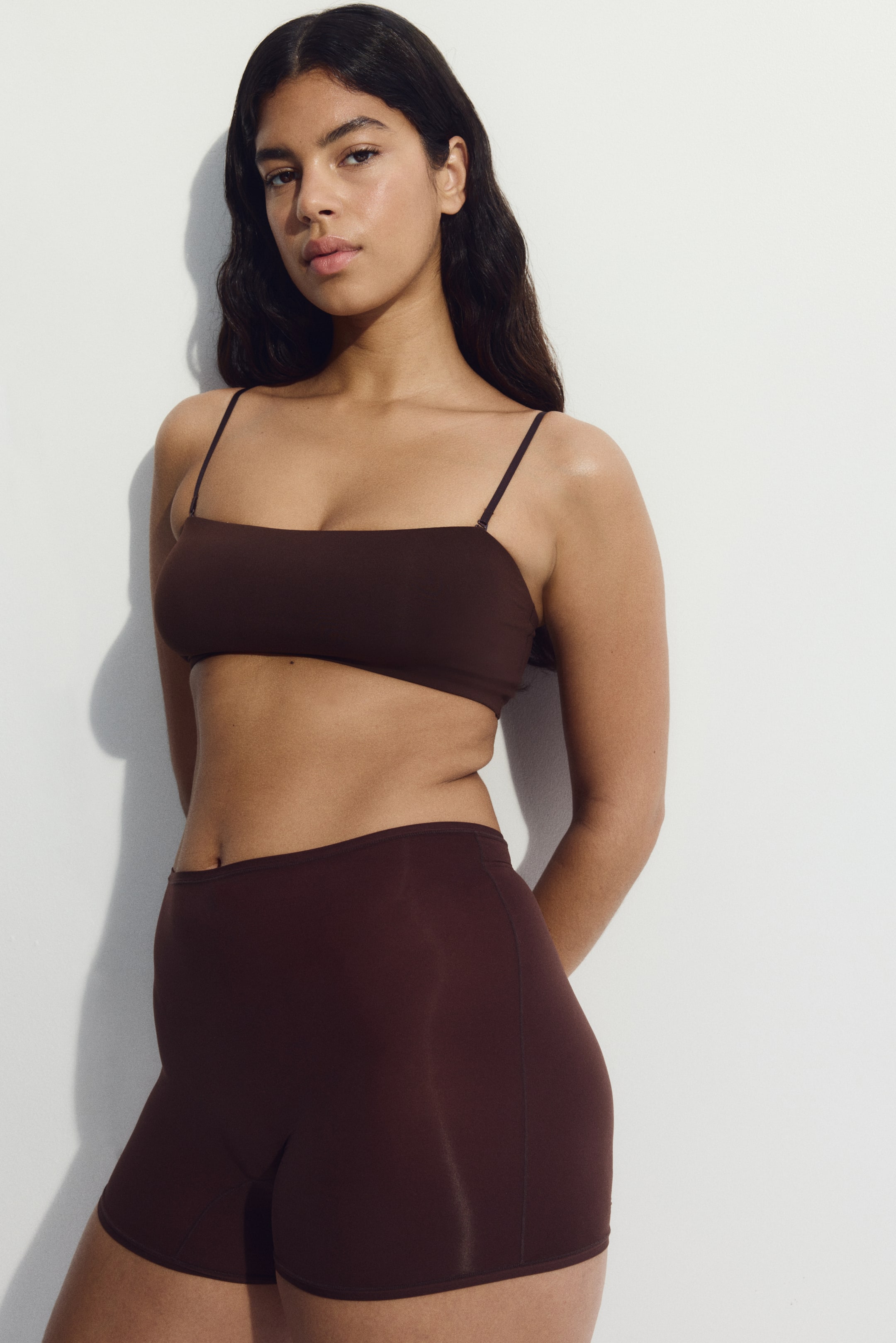 Visa större bild: En brun mikrofiber bandeau-bh med avtagbara, justerbara axelband bärs av modellen. Den är slät, utan synliga fästen, och kombineras med matchande bruna shorts.