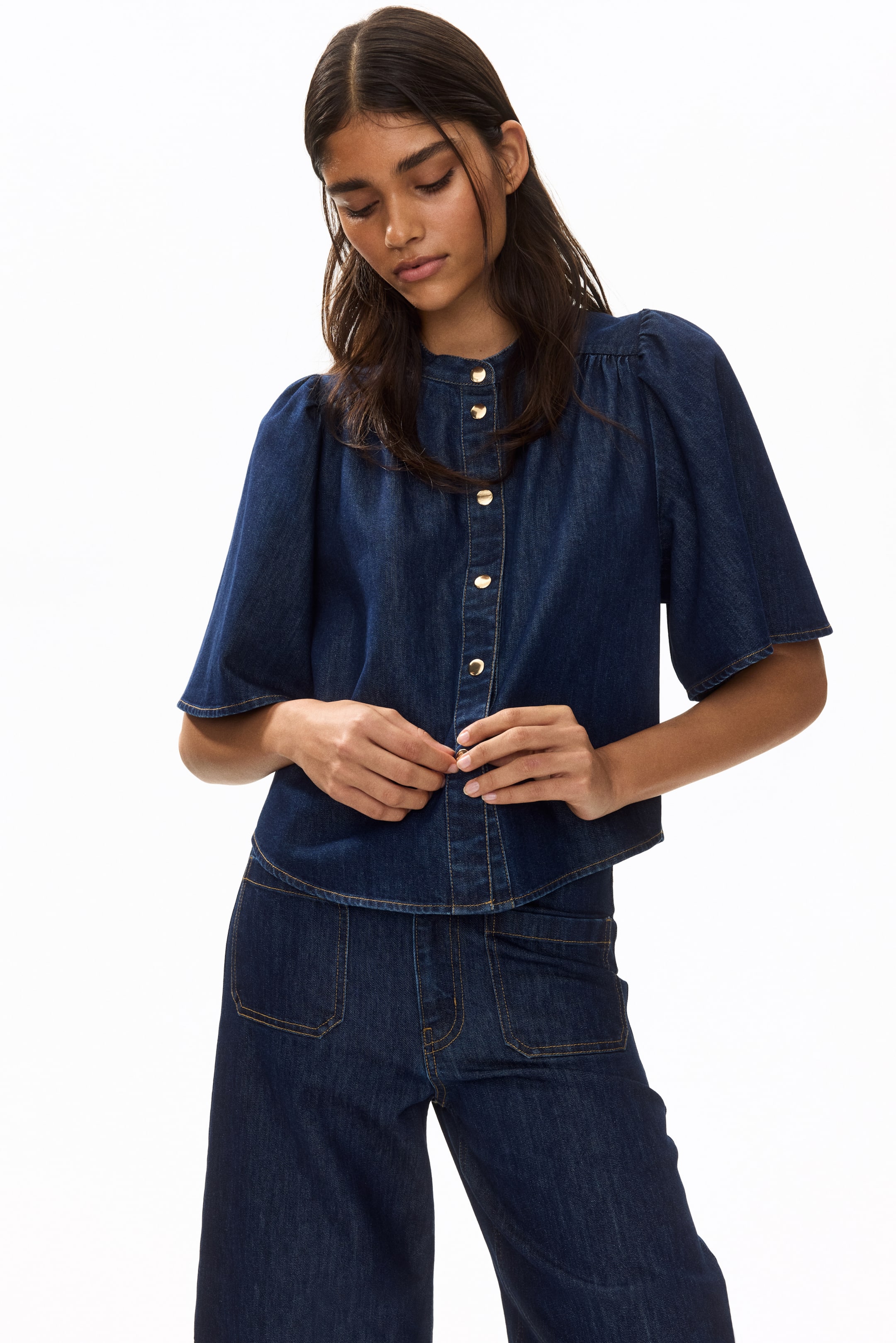 View larger image: The model is wearing an A-line blouse crafted from dark denim. This blouse showcases a band collar and gold-toned press-studs along the front. It has a relaxed fit and features short, wide sleeves adorned with gathers at the shoulders. The model is seen adjusting the front press-studs while pairing the blouse with matching dark denim trousers.