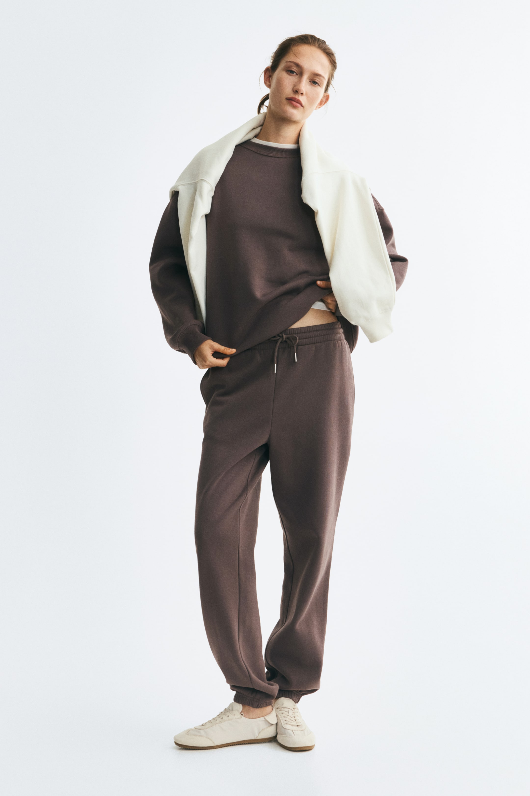 View larger image: The model is wearing track pants in a dark brown cotton-blend sweat fabric, featuring a high waist with a drawstring and elasticated hems. The pants are styled with a matching crew-neck sweatshirt and cream sneakers.