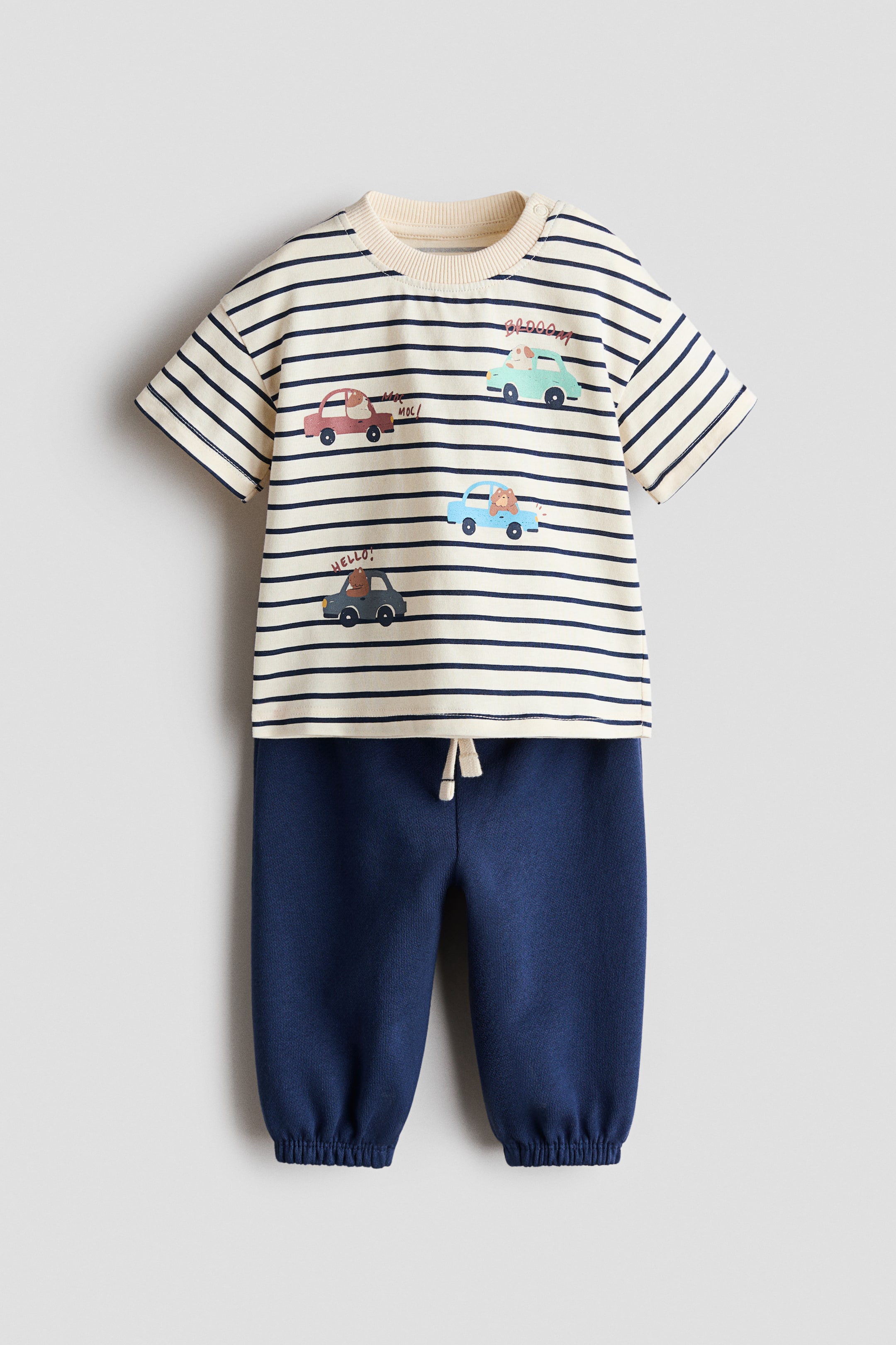 Agrandir l'image: Un ensemble pour bébé en deux pièces est présenté de face, composé d'un t-shirt à manches courtes rayé crème et bleu marine avec un col rond côtelé, et d'un pantalon jogger bleu marine. Le t-shirt affiche des illustrations de voitures avec des personnages animaliers et un texte ludique, tandis que le pantalon jogger présente une taille réglable avec cordon de serrage et des poignets de cheville côtelés.