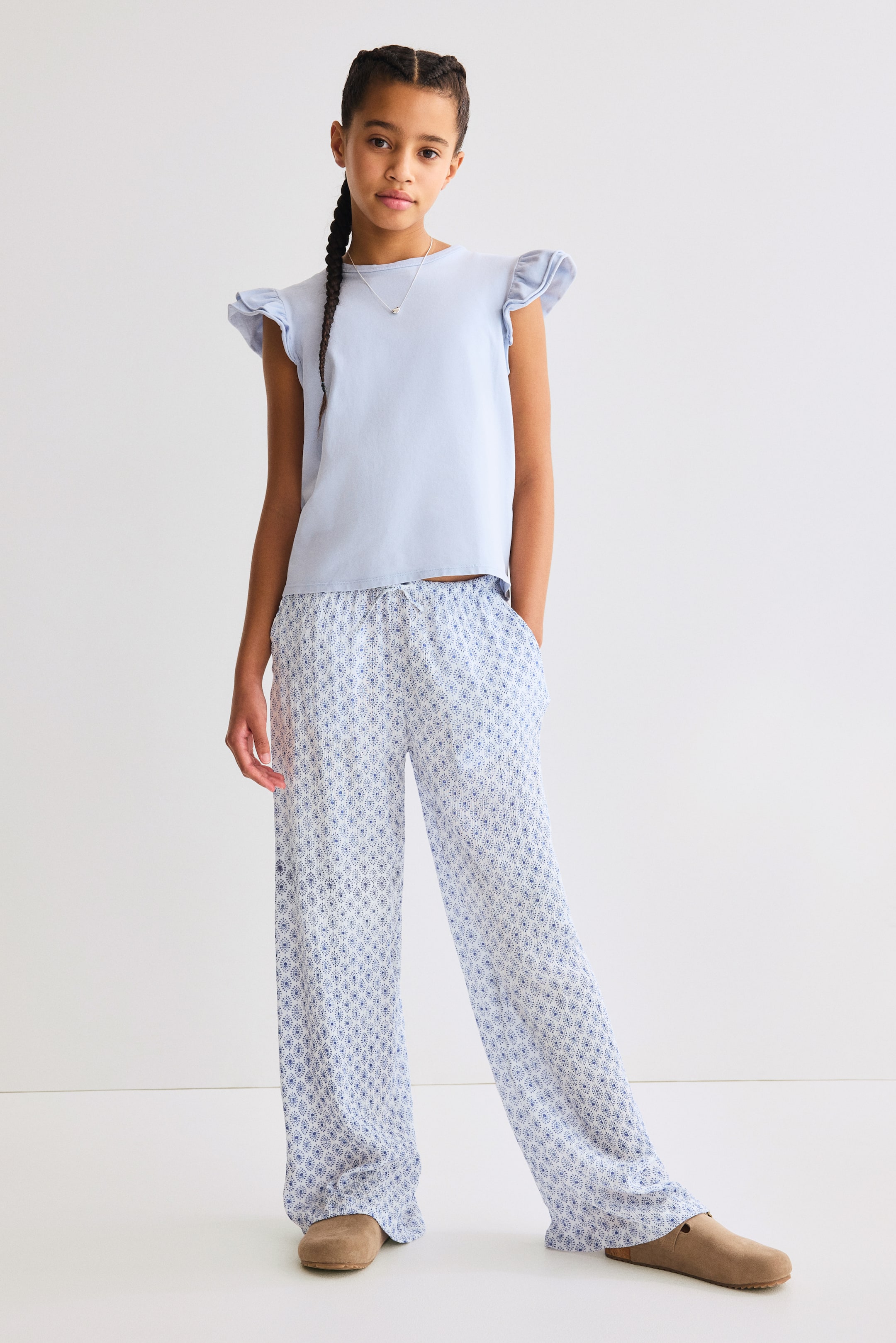 查看大圖: 淺藍色短袖上衣,搭配荷葉邊袖口,搭配寬腿褲。褲子展示了白色和藍色的幾何圖案,配有彈性抽繩腰帶和可見的側口袋。