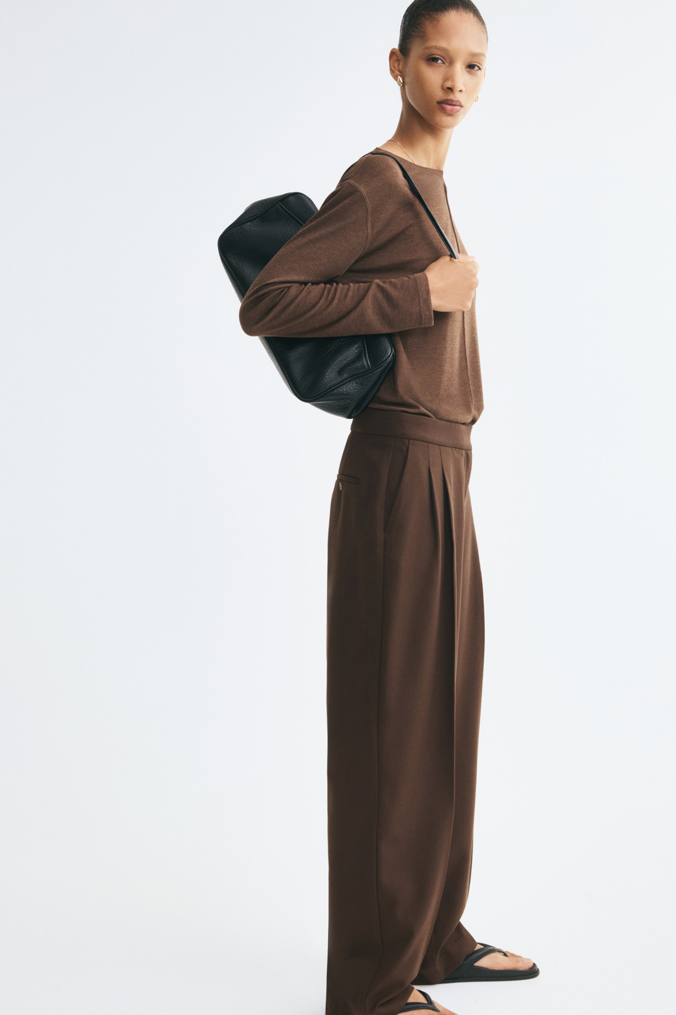 View larger image: These loose-fit, tailored twill pants in dark brown are shown from a side-profile view, featuring a high waist and a relaxed barrel-leg silhouette. The pants display sharp front pleats, creases down the legs, and diagonal side pockets, styled with a matching brown long-sleeve top.