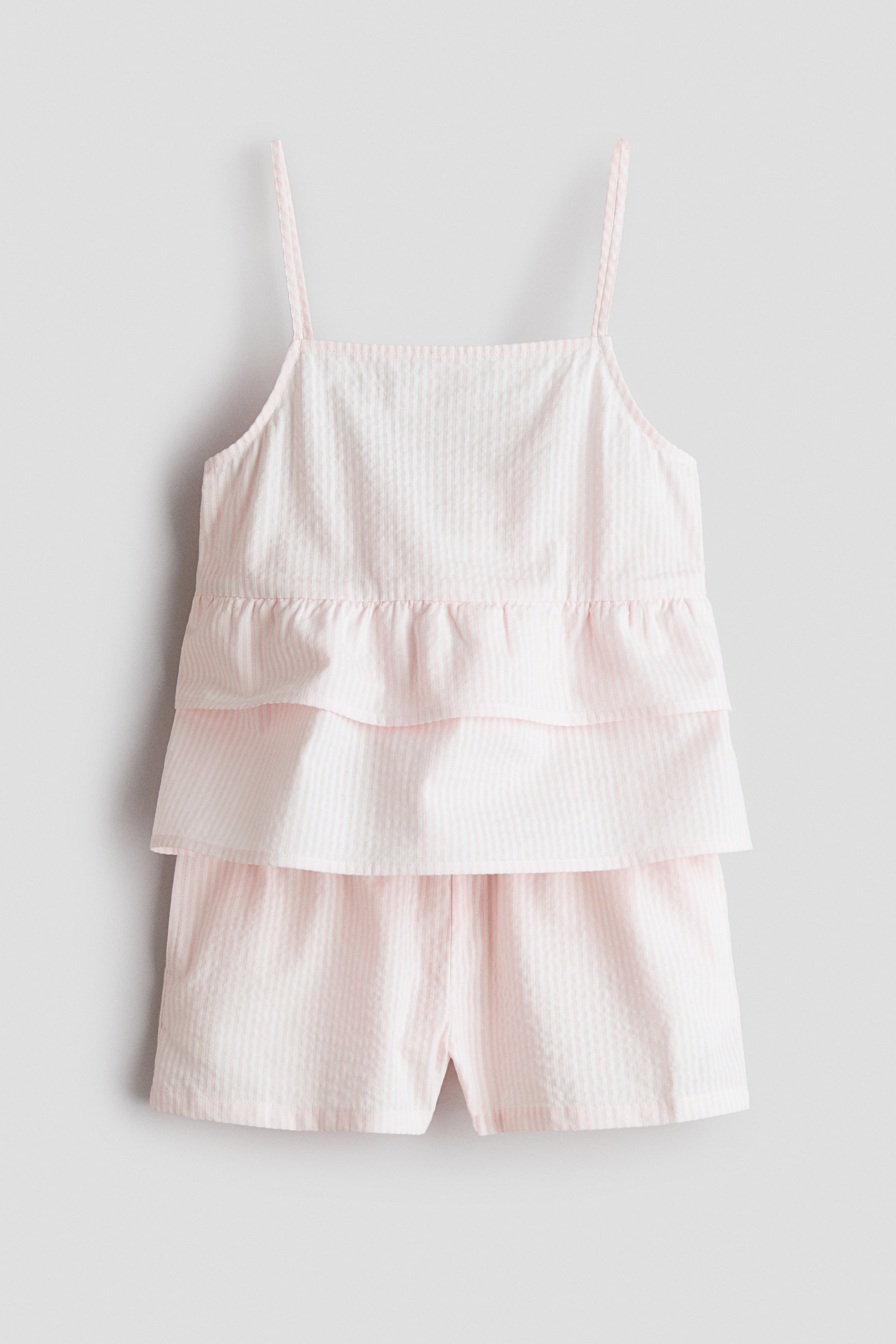 View larger image: A light pink and white striped seersucker two-piece pajama set is showcased front-facing. The camisole top features thin spaghetti straps and two ruffled tiers, while the matching shorts display a relaxed fit.
