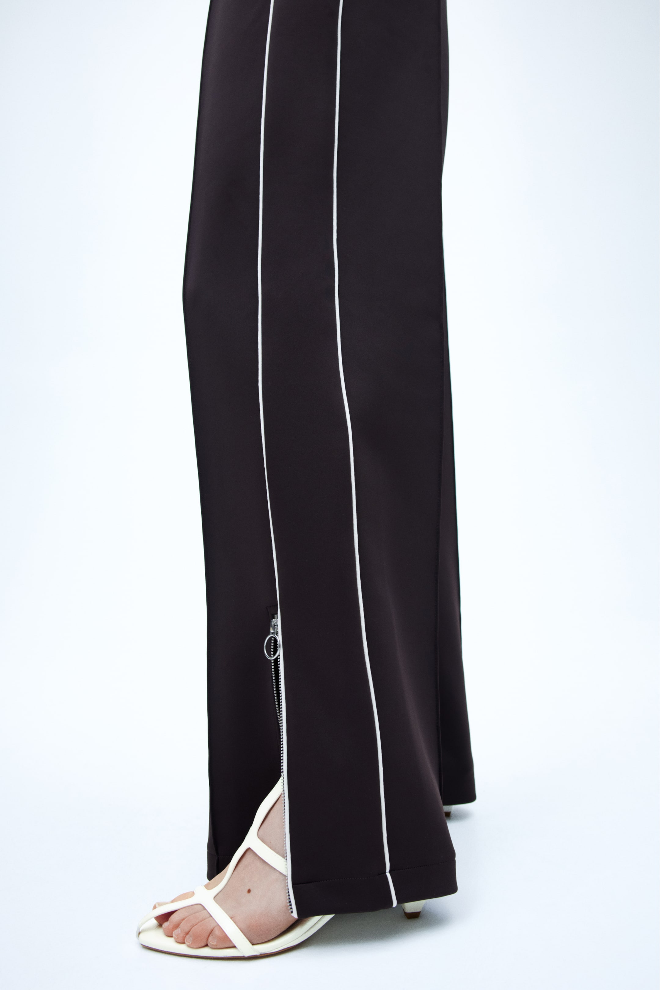 Ampliar la imagen: Pantalones marrón oscuro con una franja blanca que recorre cada costura lateral. Un cierre plateado se extiende hacia arriba desde el dobladillo en el lado exterior de una pierna, que está parcialmente abierto y revela una sandalia blanca.