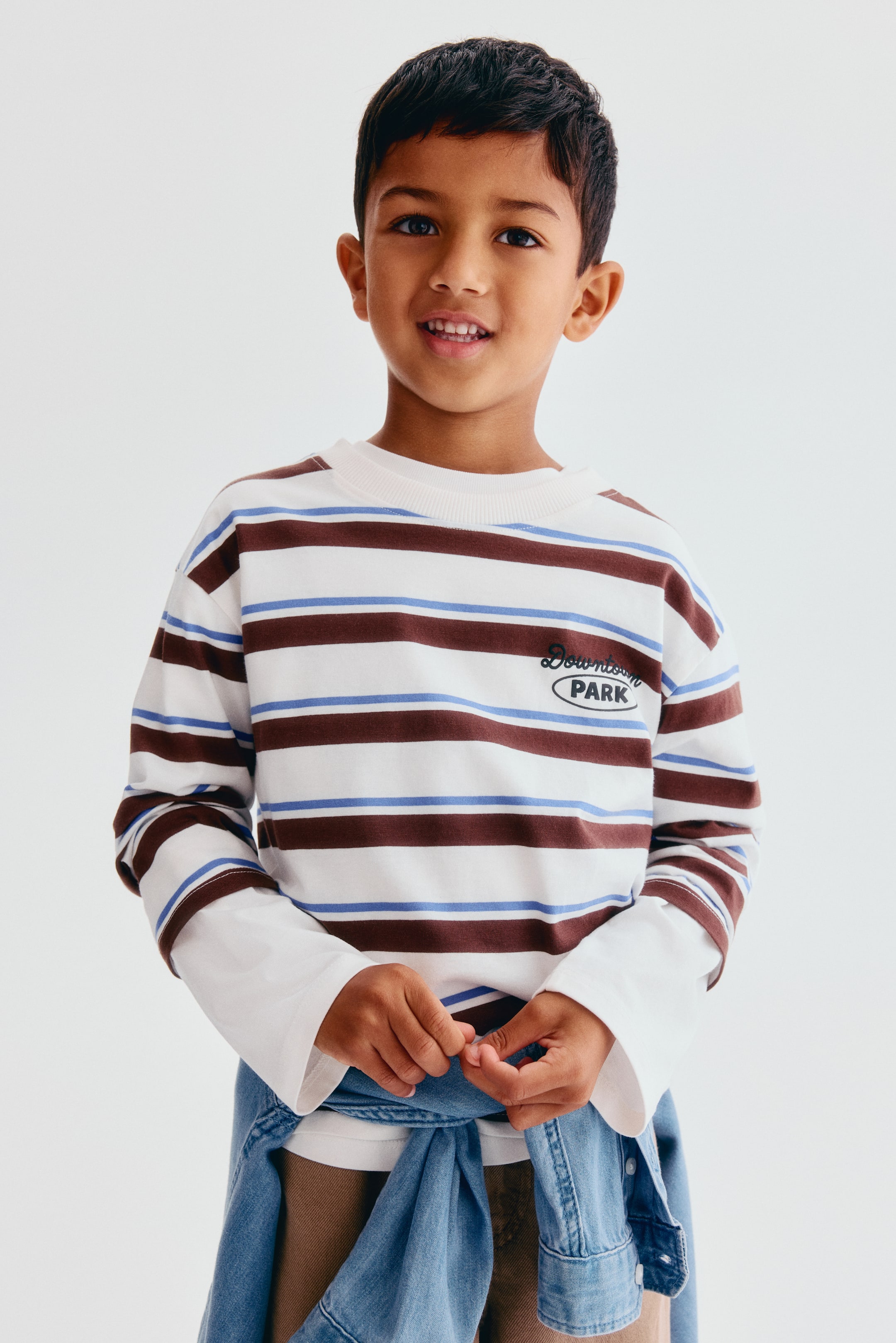Grotere afbeelding bekijken: Een T-shirt van katoenen jersey met lange mouwen is van voren afgebeeld, met horizontale bruine en lichtblauwe strepen op een witte basis, een geribde ronde hals, verlaagde schouders en een geprinte 'Downtown PARK'-opdruk op de borst. Het T-shirt is gedragen over een wit shirt met lange mouwen en gestyled met een lichtblauw denim hemd dat rond de taille is geknoopt.