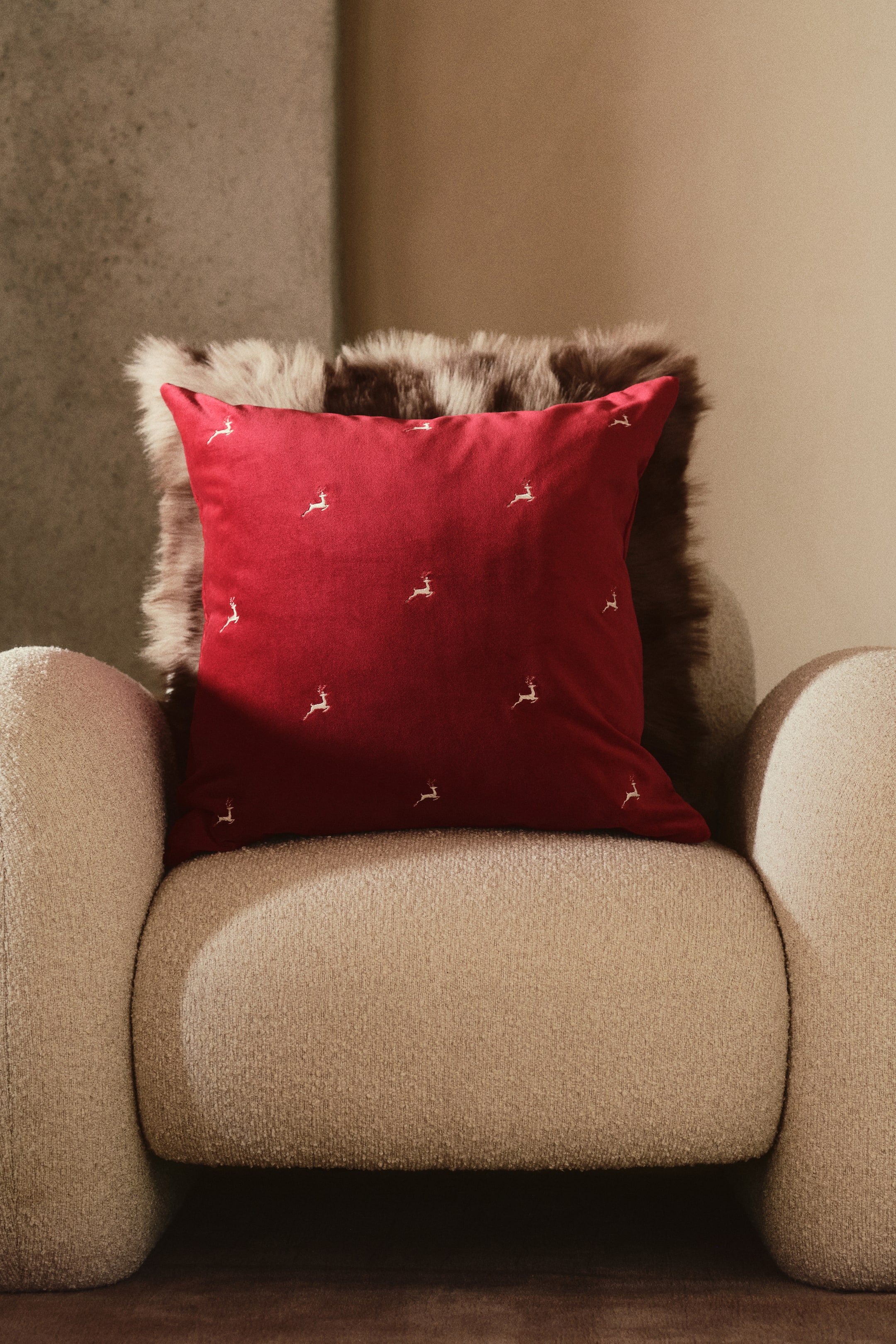 Ampliar la imagen: Una funda de cojín cuadrada de terciopelo de algodón rojo descansa de frente sobre un sillón beige de bouclé, mostrando pequeños motivos de renos bordados y dispersos, cosidos con hilo brillante. El cojín rojo está colocado directamente al frente de una almohada más grande, esponjosa y marrón de piel sintética.