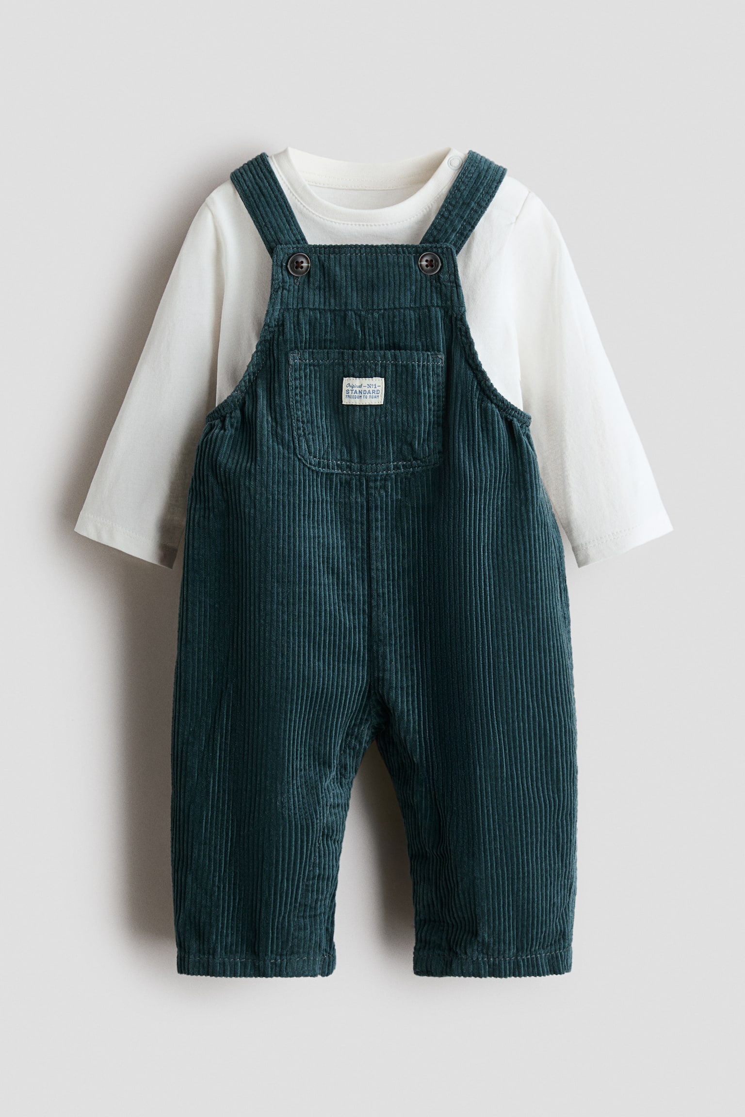 A front-facing baby outfit features dark teal corduroy overalls layered over a plain white long-sleeve T-shirt. The overalls feature button-fastened shoulder straps, a chest patch pocket with a small white label, and straight-cut legs.