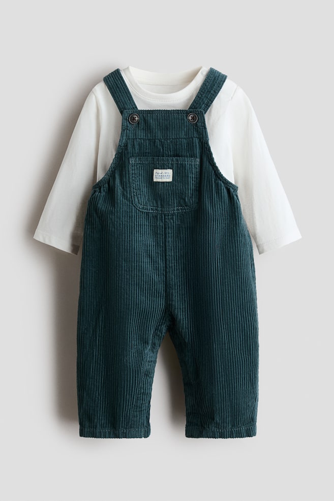 A front-facing baby outfit is featuring dark teal corduroy overalls layered over a plain white long-sleeve t-shirt. The overalls are displaying button-fastened shoulder straps, a chest patch pocket with a small white label, and straight-cut legs. - 1