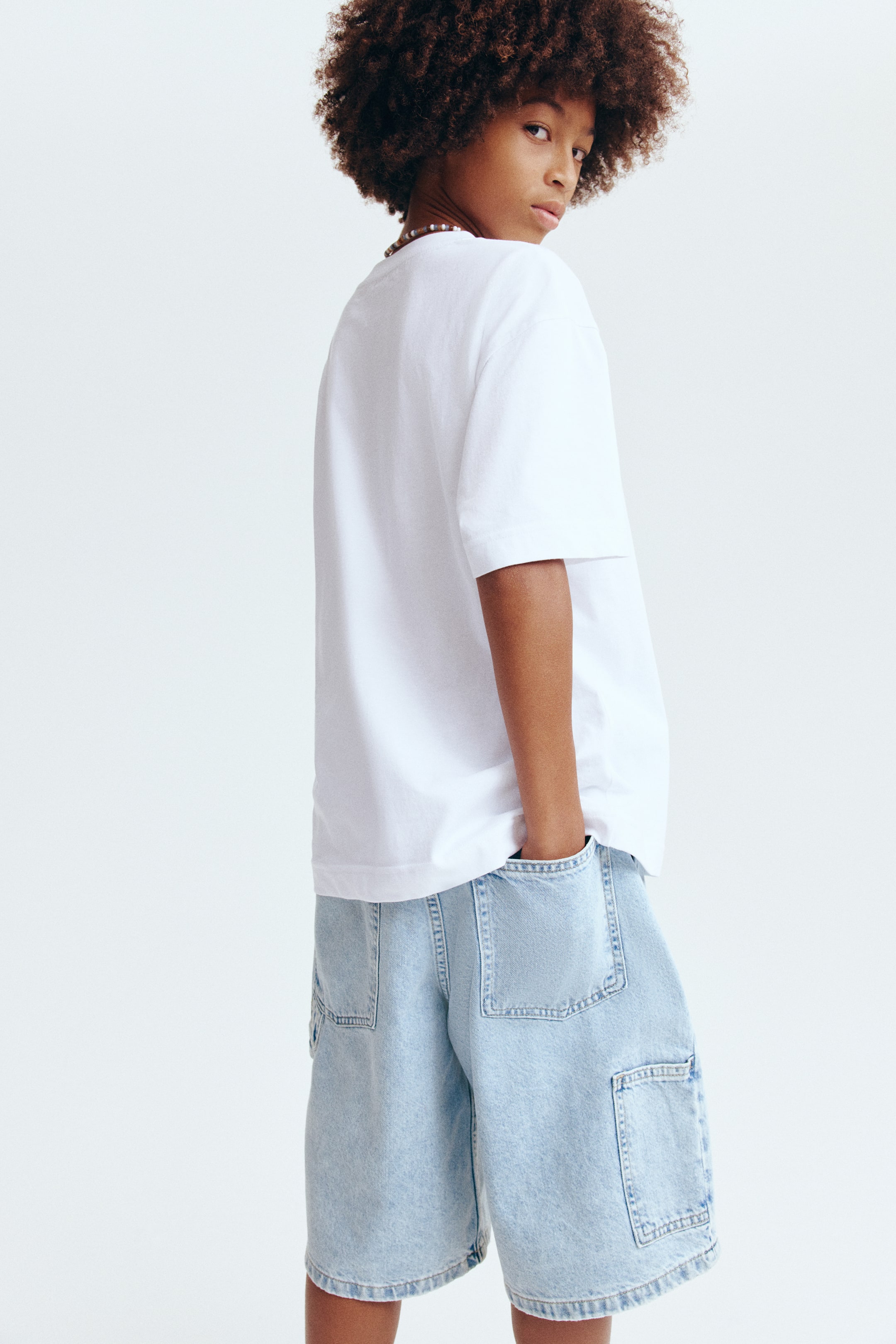 View larger image: A white crew neck t-shirt with short sleeves and light blue denim cargo shorts are being showcased from a back-side angle. The t-shirt features a relaxed fit, while the shorts display large patch pockets on the back and a utility pocket on the side, highlighted by visible stitching details.