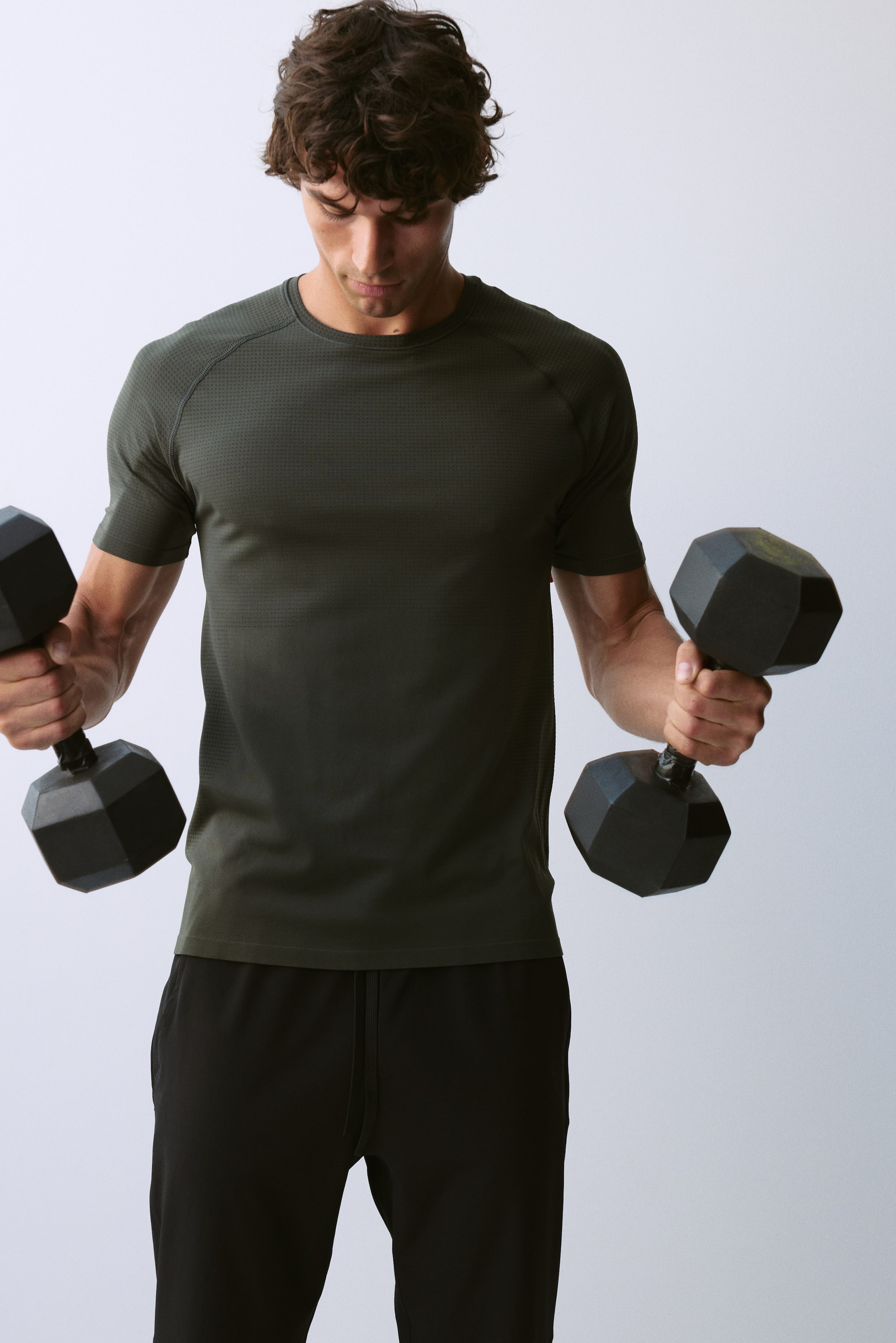 View larger image: The model is wearing an olive green crew-neck athletic t-shirt with raglan short sleeves and a textured knit pattern. It is styled with black athletic pants, while the model holds a black hexagonal dumbbell in each hand.
