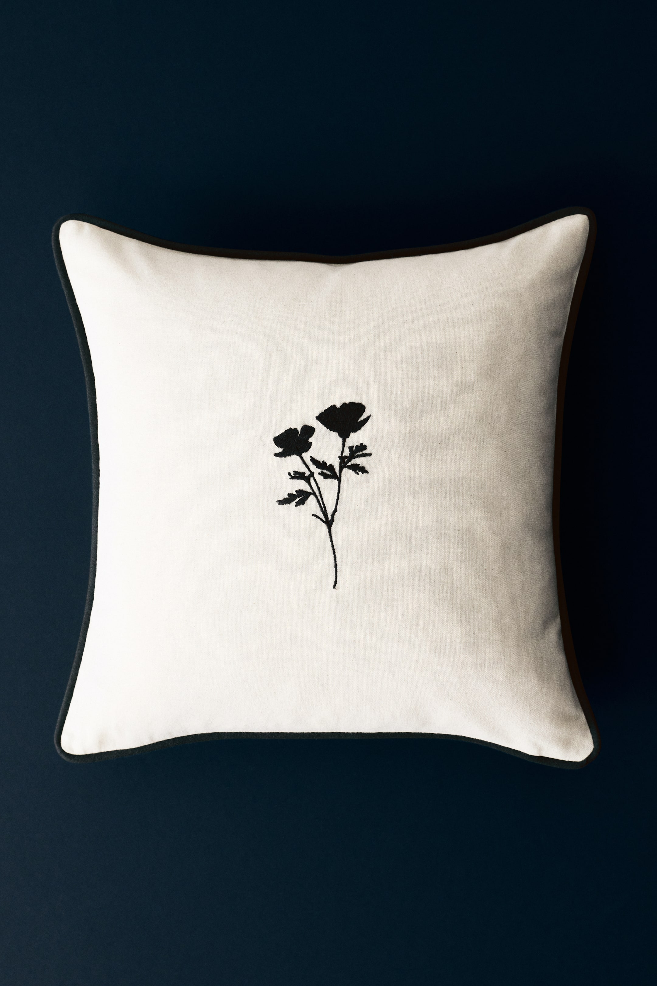 Größeres Bild ansehen: Ein quadratischer Kissenbezug aus cremefarbenem Baumwoll-Canvas ist von vorne zu sehen, mit einem schwarzen, gestickten Blumenmotiv in der Mitte und kontrastierender dunkler Paspelierung entlang der Kanten.
